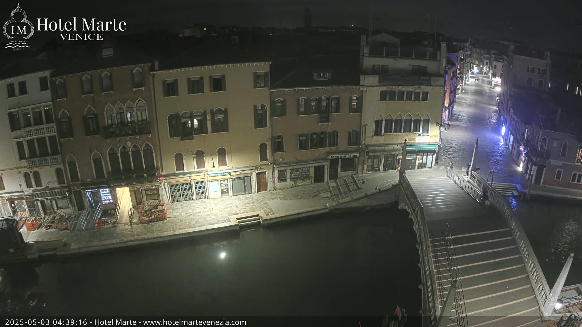 A nighttime view of a canal in Venice, showing buildings lining the waterway, a bridge with steps, and flooded streets.