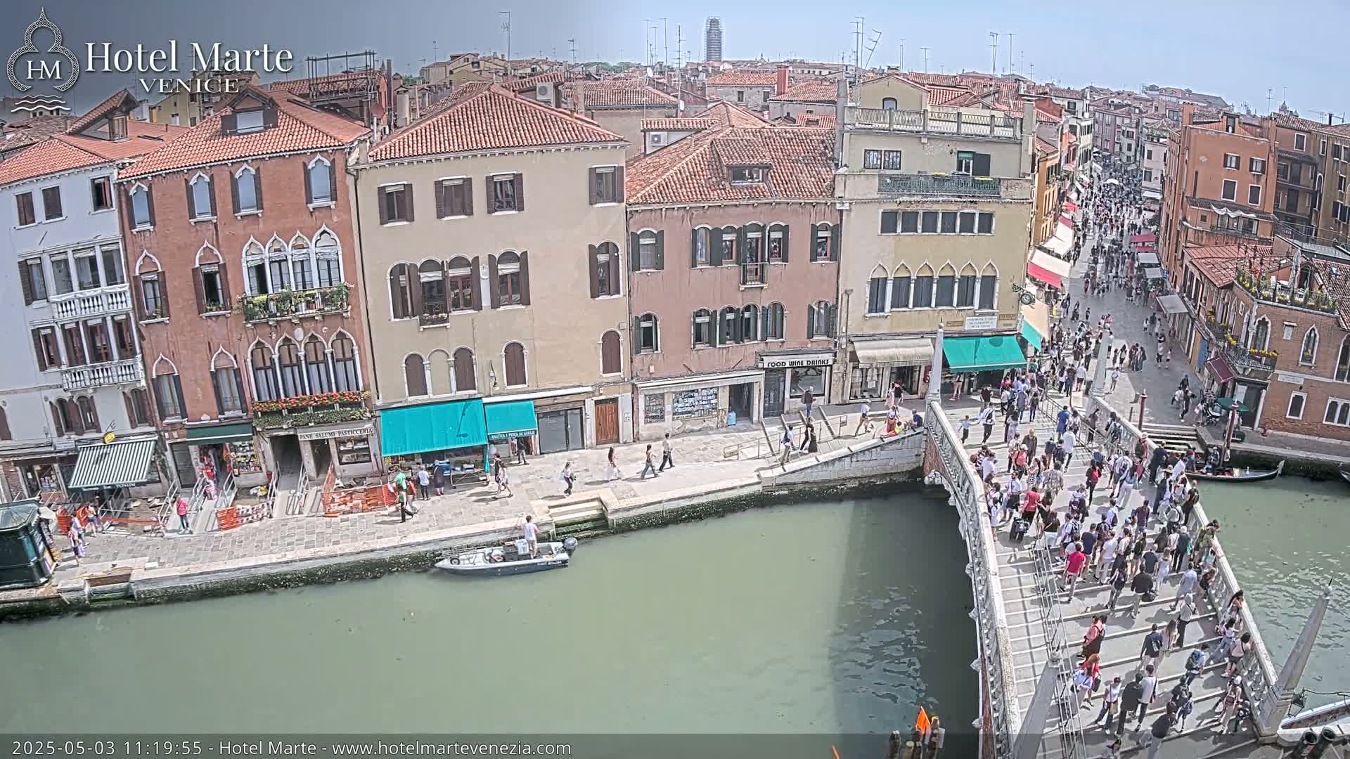 A crowded footbridge crosses a canal in Venice on a sunny day, with many people walking across it and along the canal's edge, and numerous buildings lining the waterway.