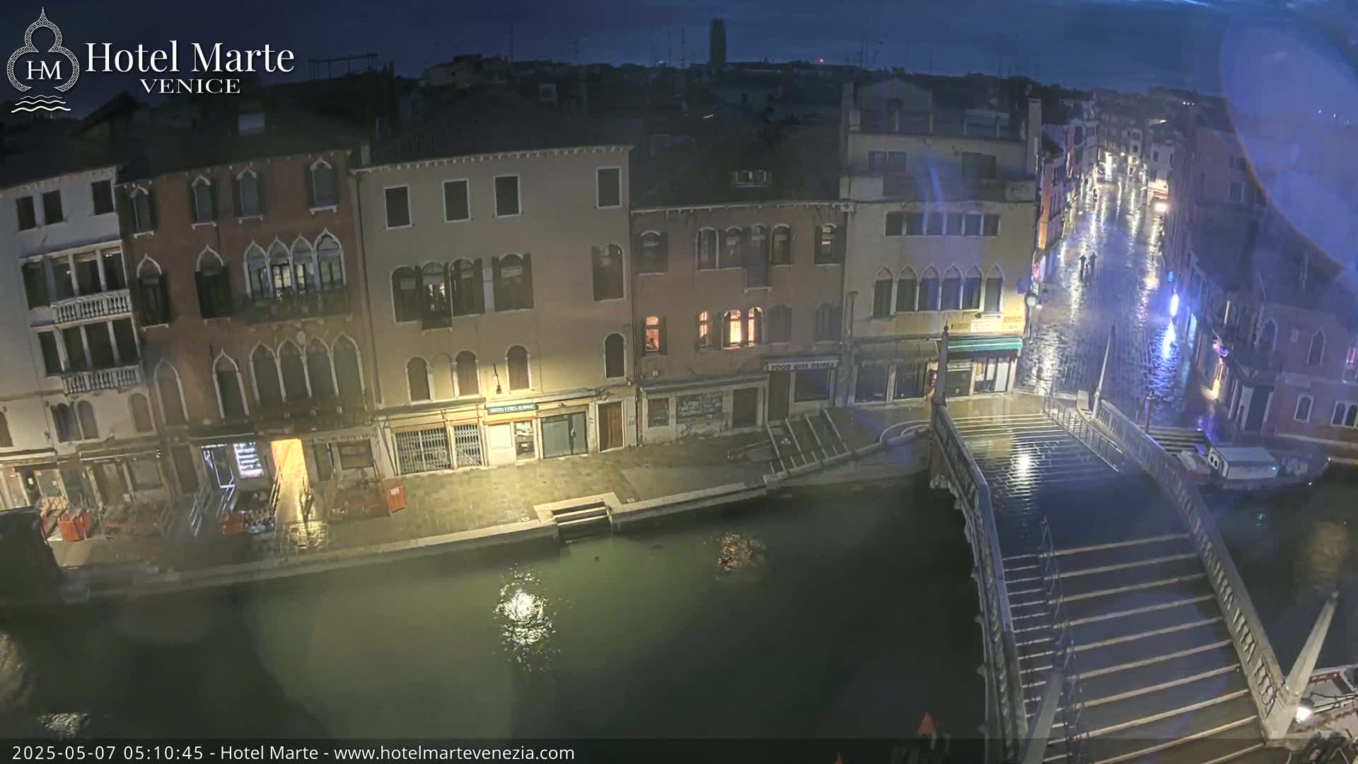 A nighttime view of a canal in Venice, showing several buildings along the waterway and a wet bridge under a dark sky.