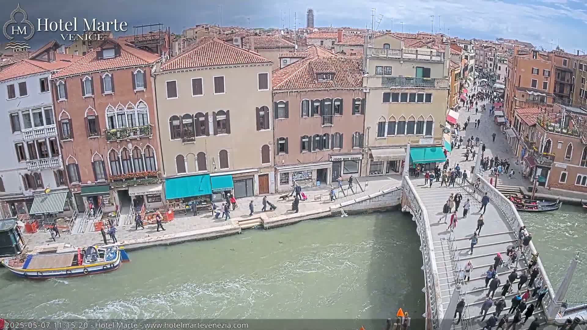 A sunny day in Venice shows a canal with a stone bridge crowded with pedestrians, boats, and buildings lining the waterway.