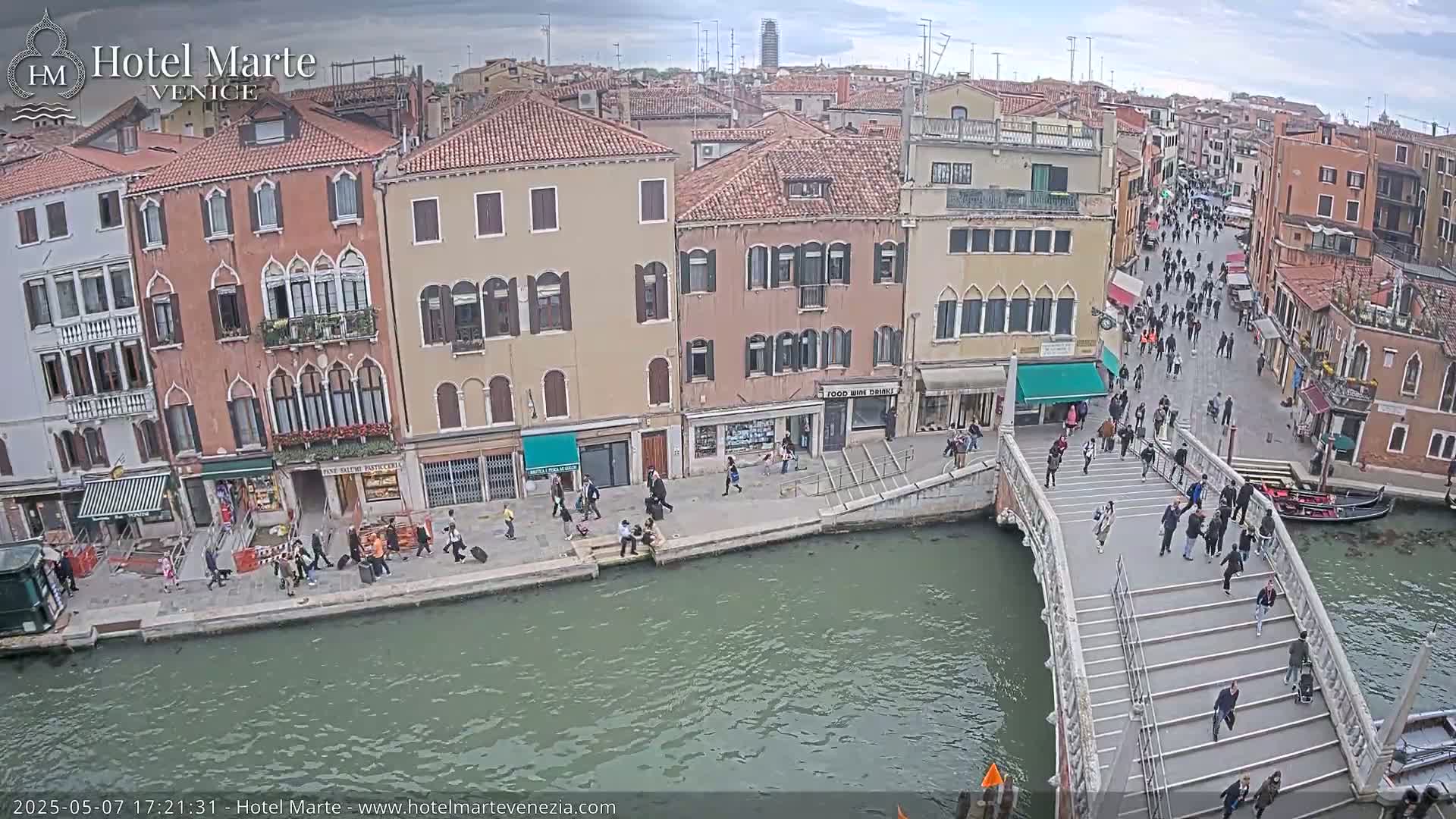 A canal in Venice, under an overcast sky, is spanned by a bridge with many pedestrians walking across it and along the canal's edge.