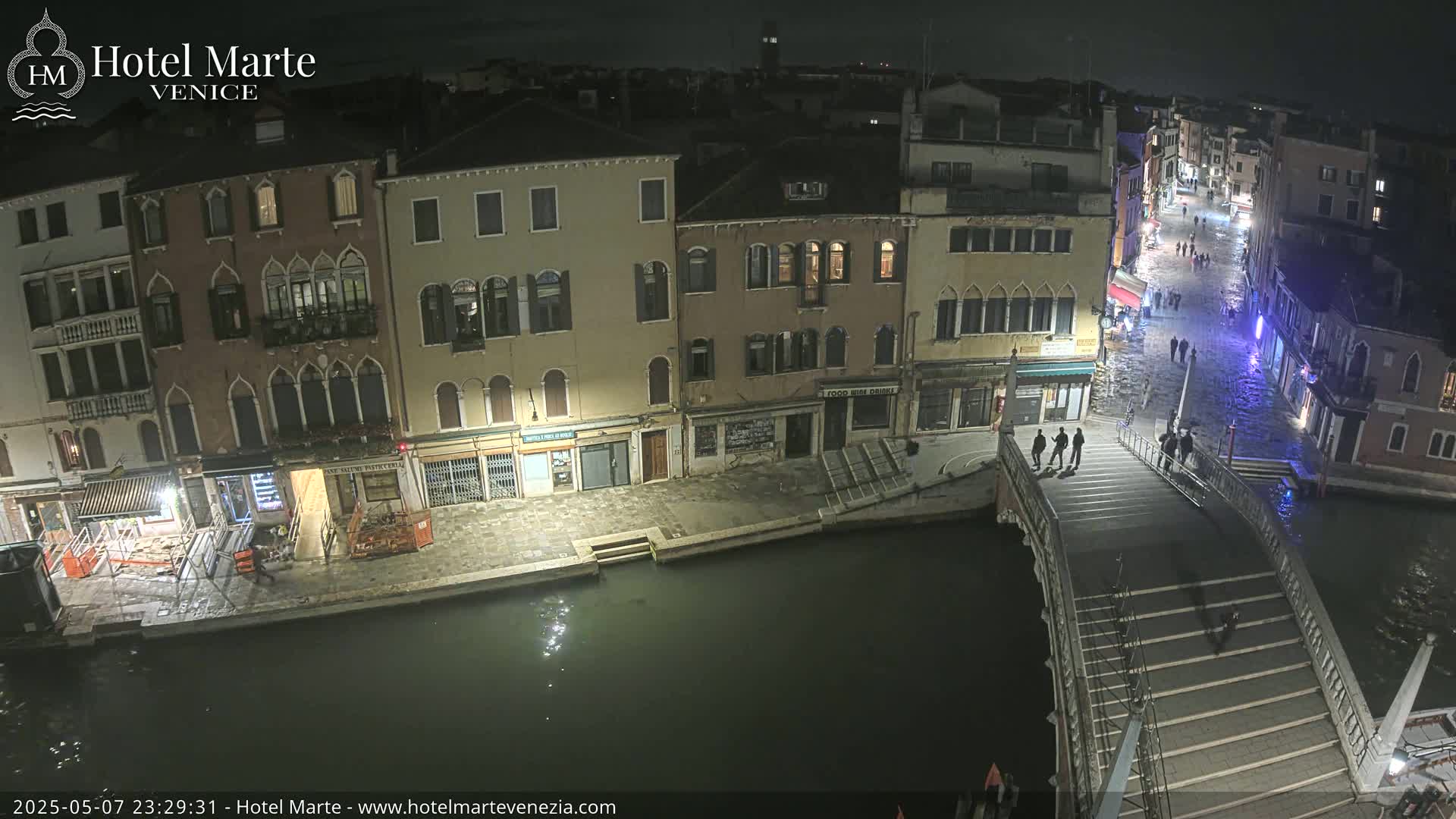 At night, a canal in Venice shows several buildings along its edge, a bridge with people walking across it, and a dark, calm waterway.