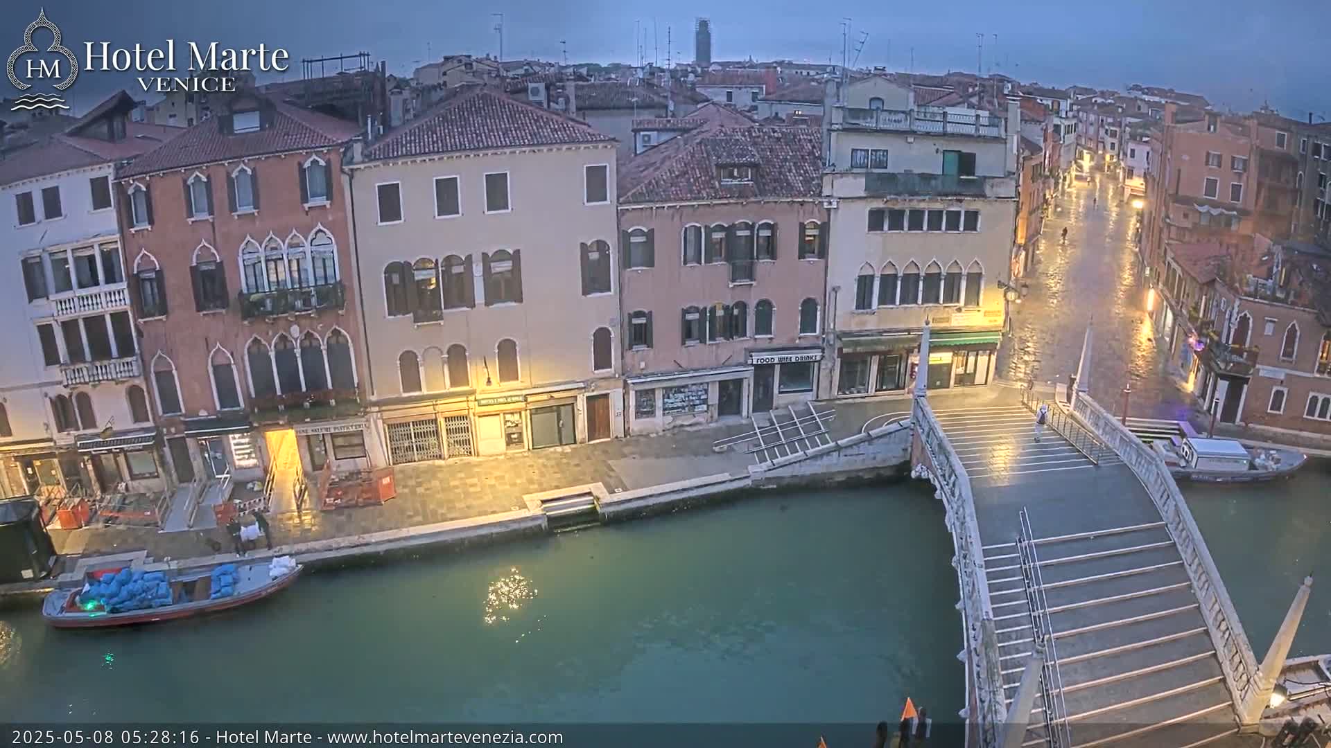 A canal in Venice, under a dark, possibly overcast sky, shows several buildings along its banks and a pedestrian bridge crossing the water.