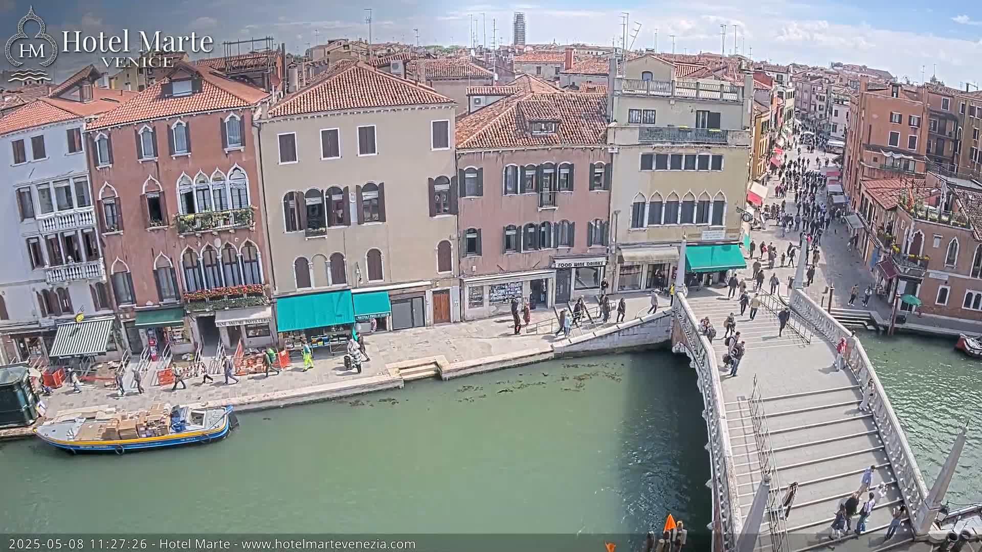 A sunny day in Venice shows a canal with a bridge crowded with pedestrians,  boats moored along the canal-side, and colorful buildings lining the waterway.