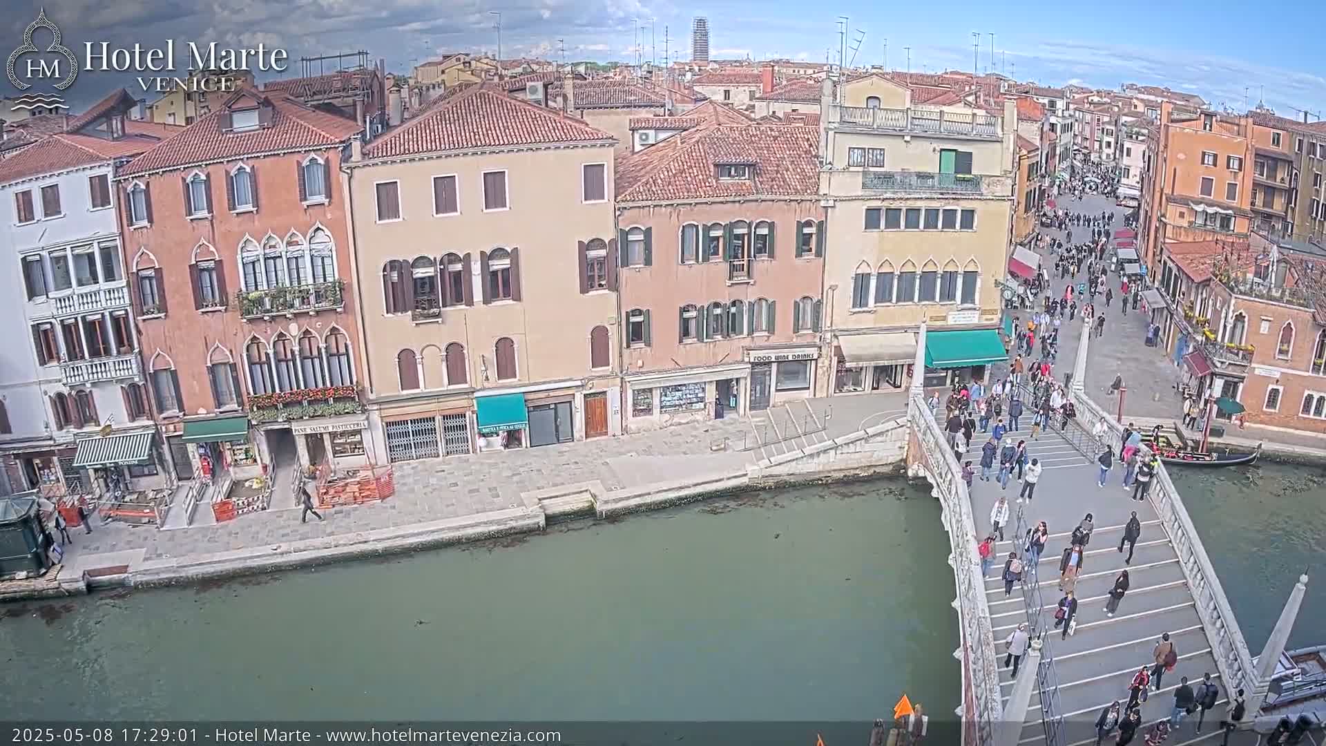 A sunny day in Venice shows many people walking across a bridge over a canal, with several multi-story buildings lining the waterway.