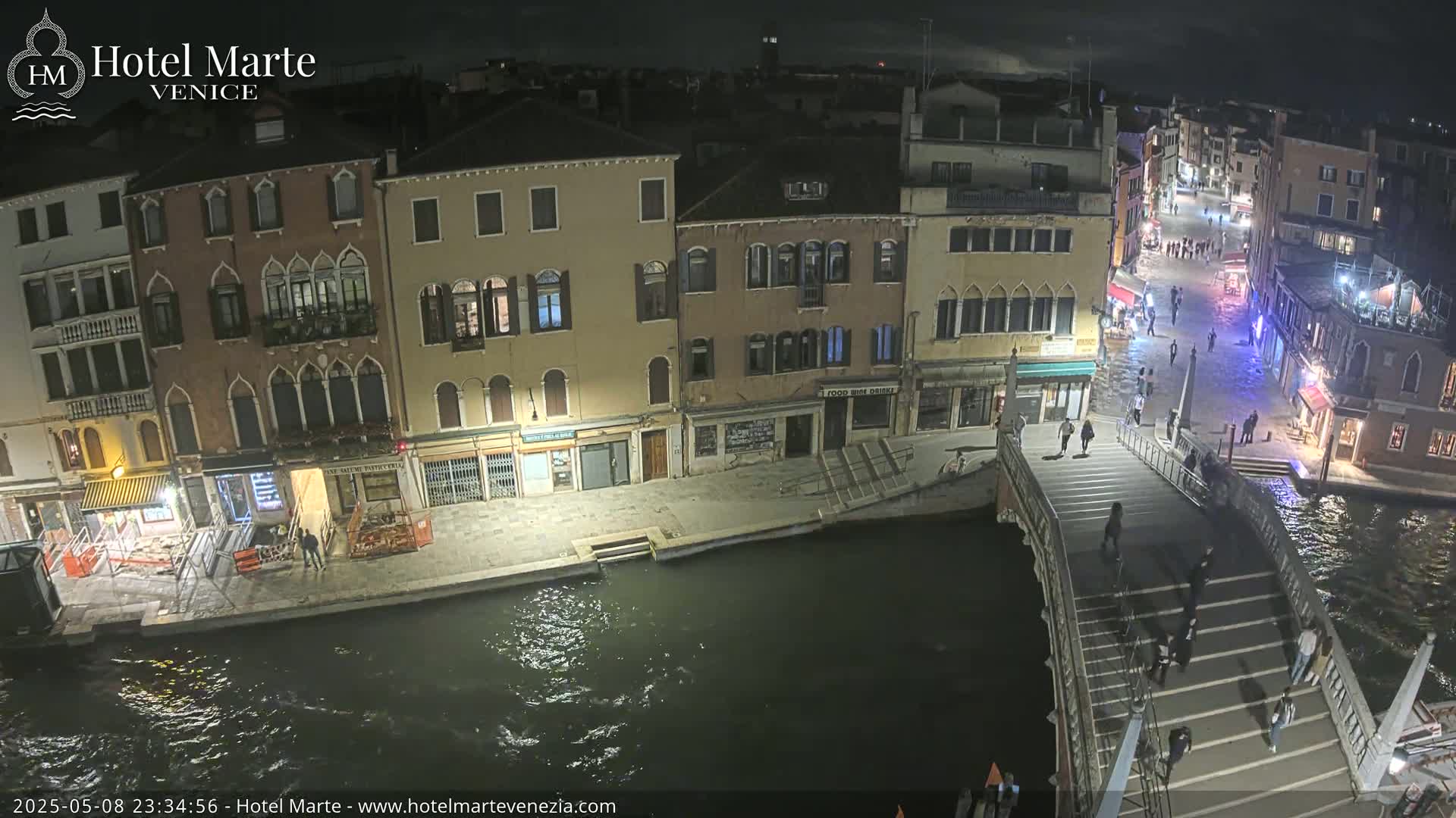 A nighttime aerial view shows a canal in Venice with several illuminated buildings lining its banks, a bridge with pedestrians crossing, and dark water.