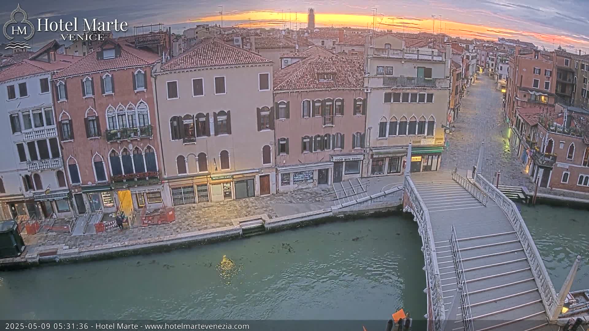 A sunrise illuminates a quiet Venetian canal scene, showcasing several buildings and a stone footbridge, with calm water reflecting the light.