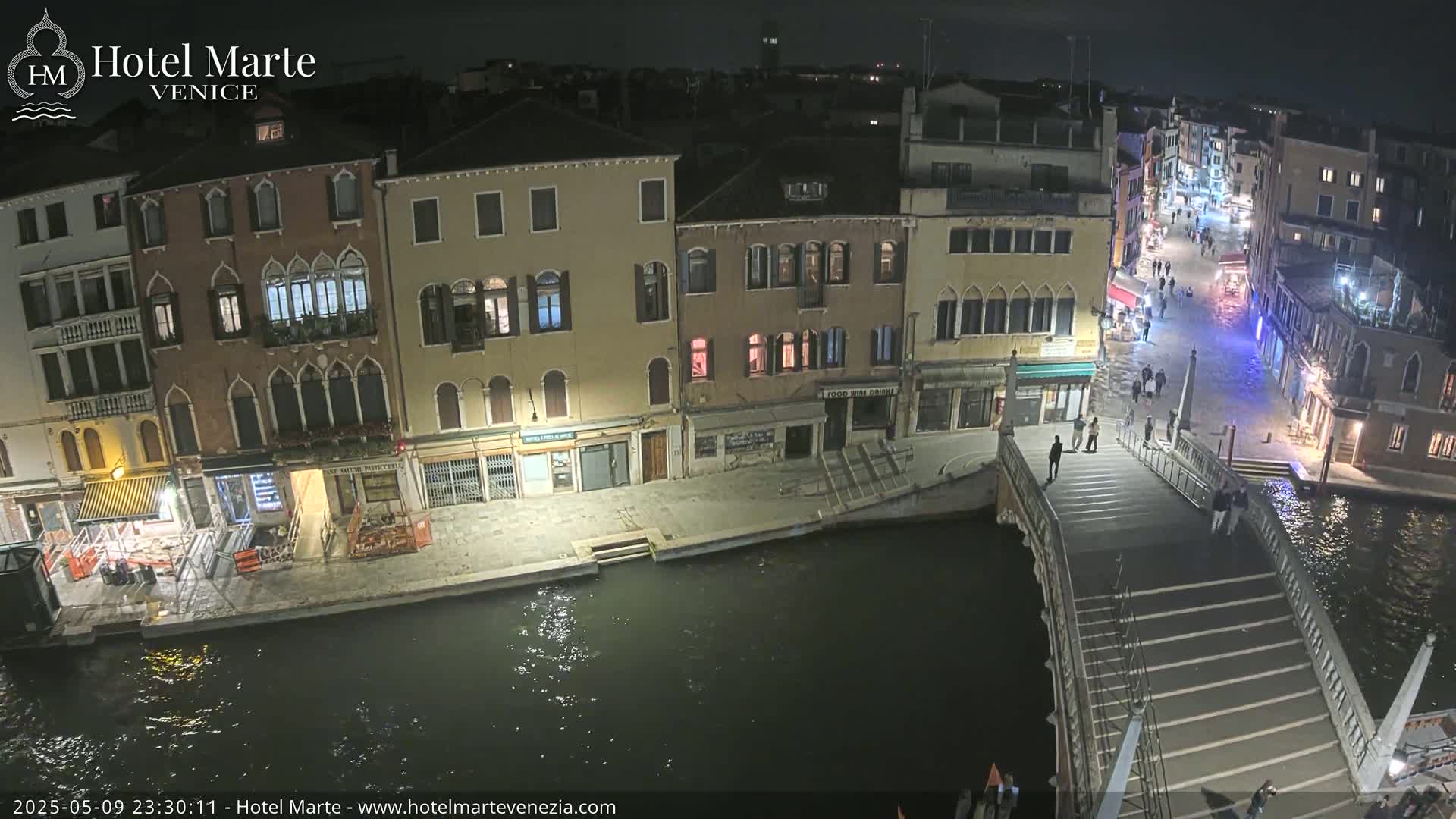 A nighttime view of a canal in Venice, showing several buildings along the waterway, a bridge with pedestrians, and dark water.