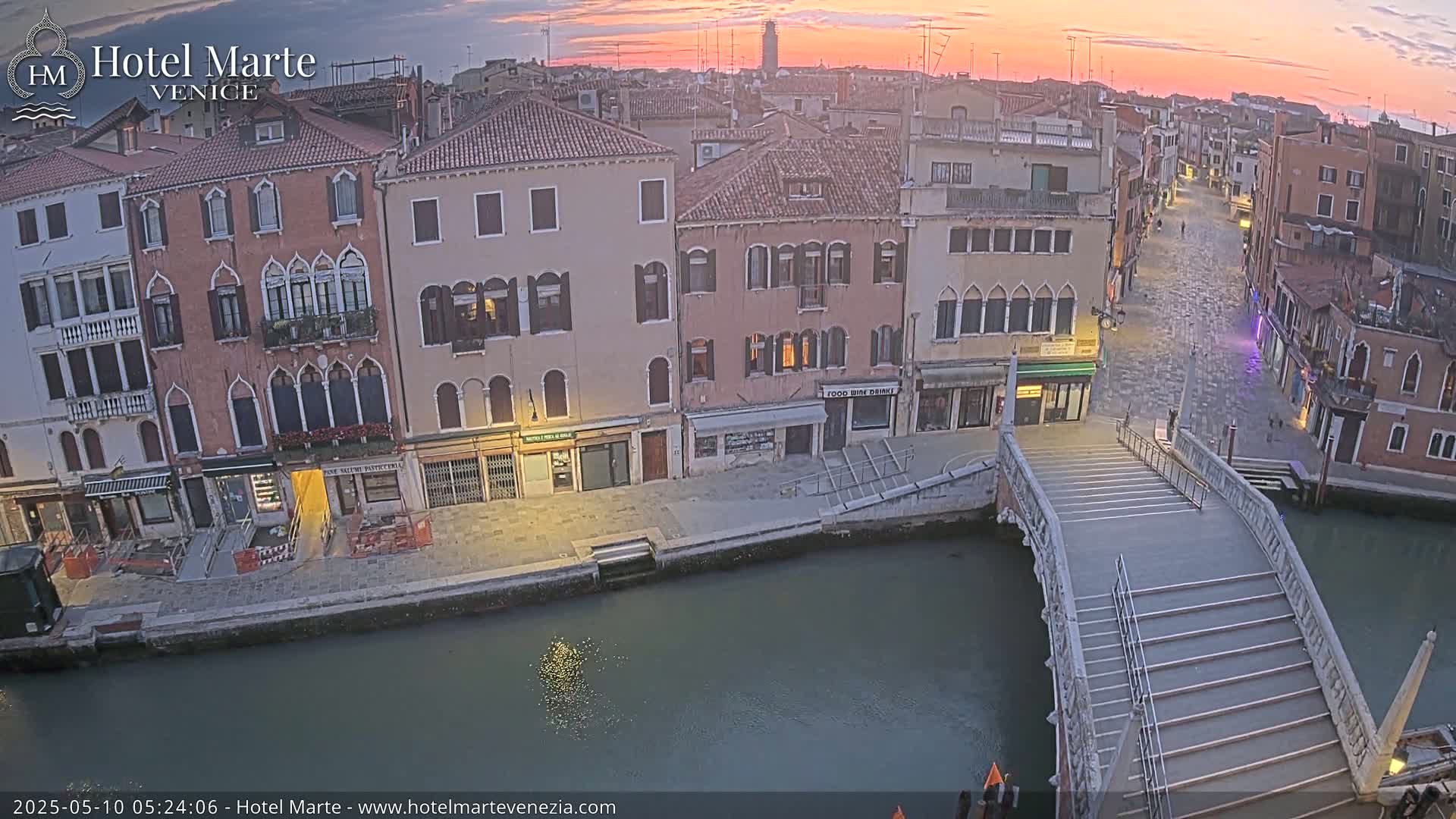 A tranquil canal scene in Venice at sunrise, showing colorful buildings lining a waterway with a stone bridge and a quiet street.