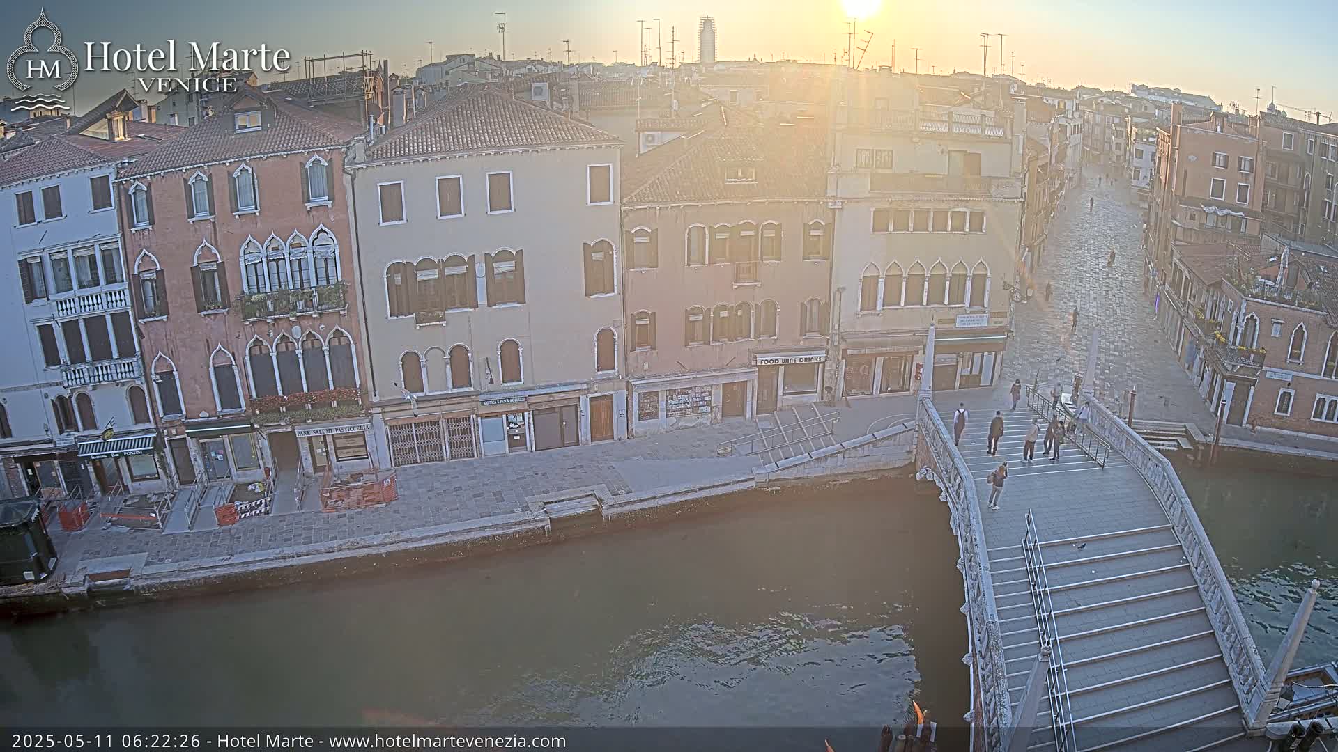 A sunrise illuminates a canal-side street in Venice, with several pedestrians walking across a stone bridge.