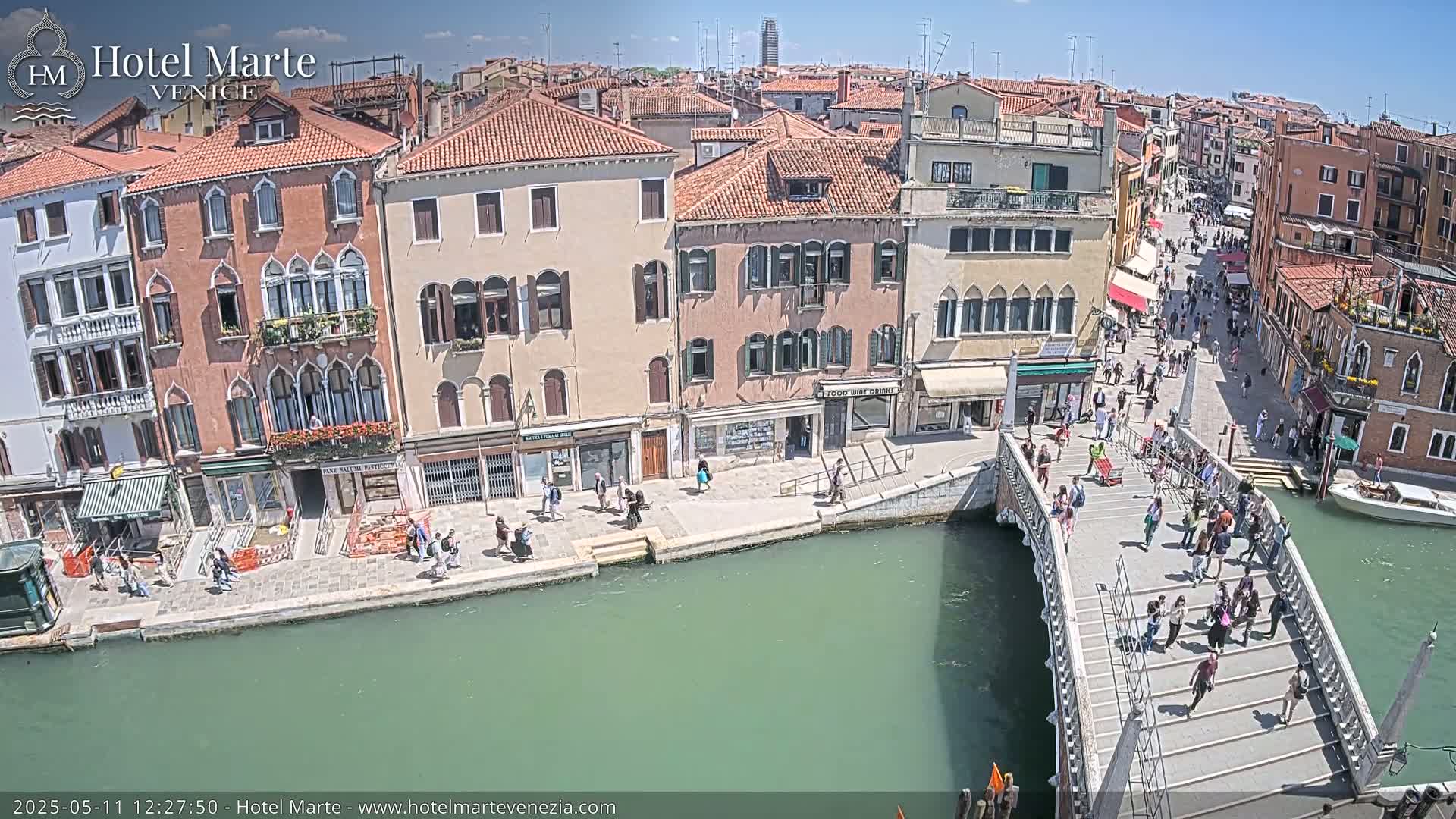 A sunny day in Venice shows a canal with a bridge crowded with people, alongside buildings and walkways bustling with activity.