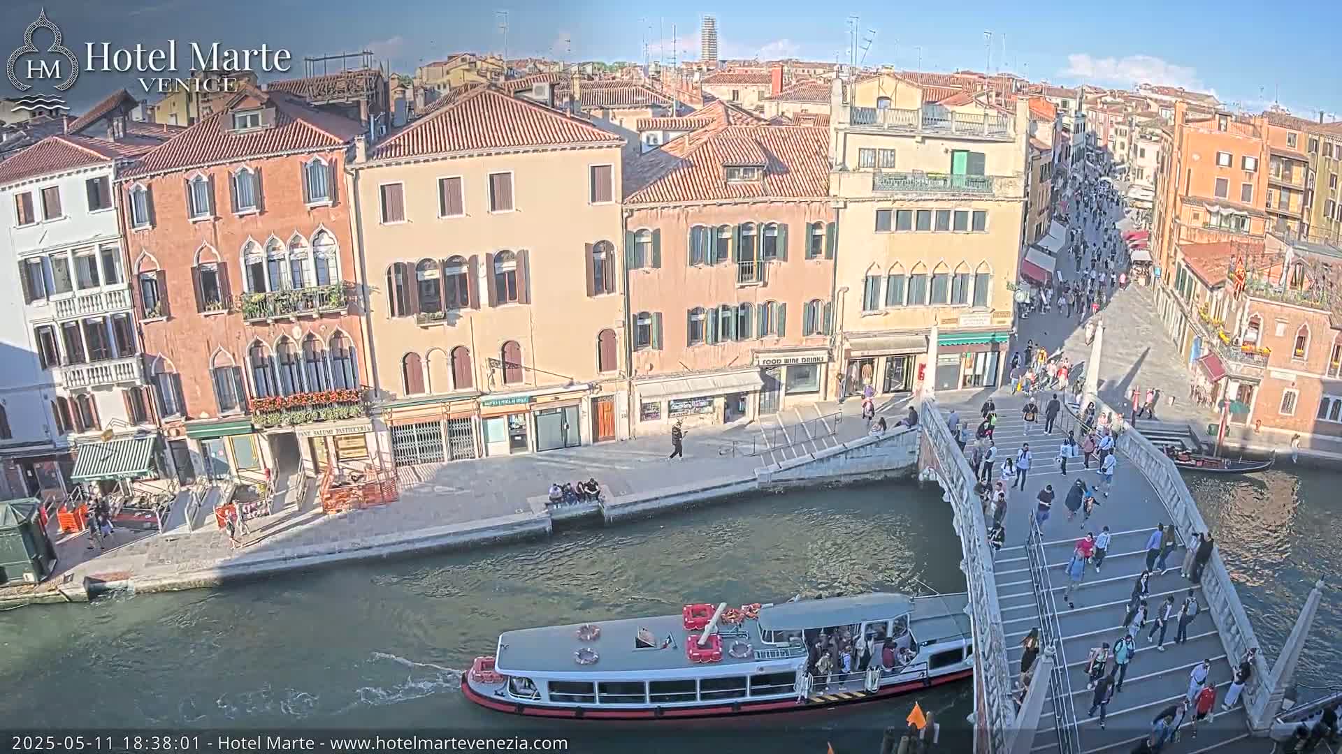 A sunny day in Venice shows a canal with a passenger boat, a bridge crowded with pedestrians, and colorful buildings lining the waterway.