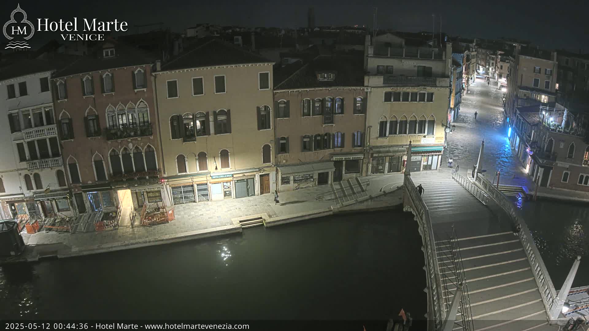 A nighttime view of a canal in Venice shows several buildings lining the waterway, a bridge with steps leading down to the water, and a few pedestrians.
