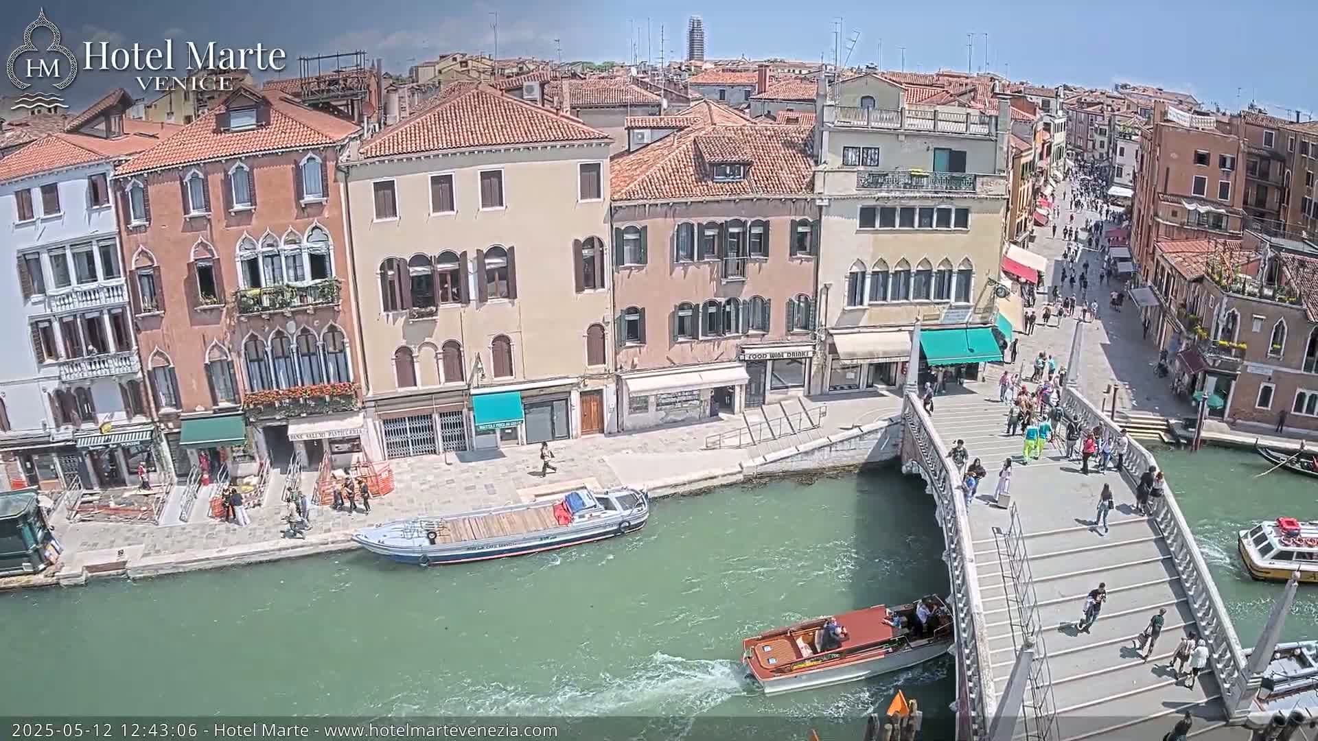 A sunny day in Venice shows a canal with several boats, a footbridge crowded with people, and colorful buildings lining the waterway.