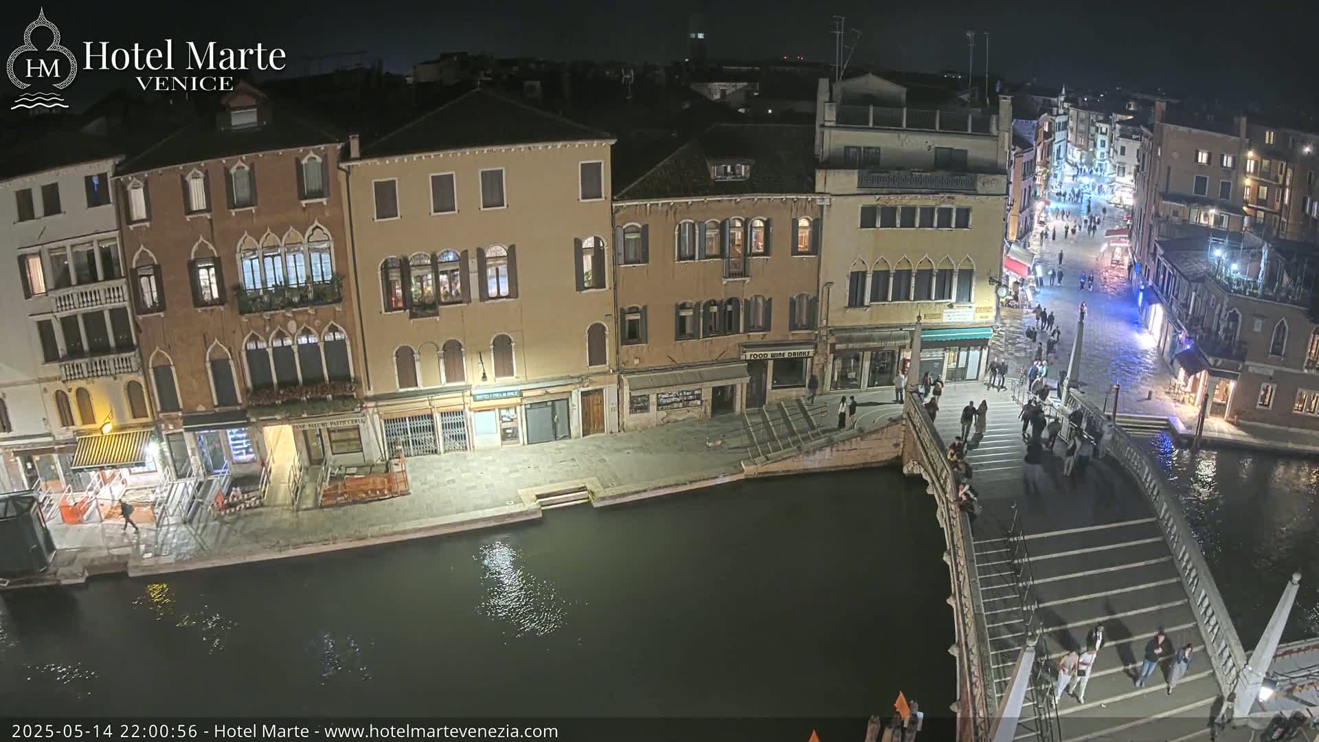 A nighttime view of a canal in Venice, showing several buildings, a bridge with people walking on it, and dark water reflecting light.