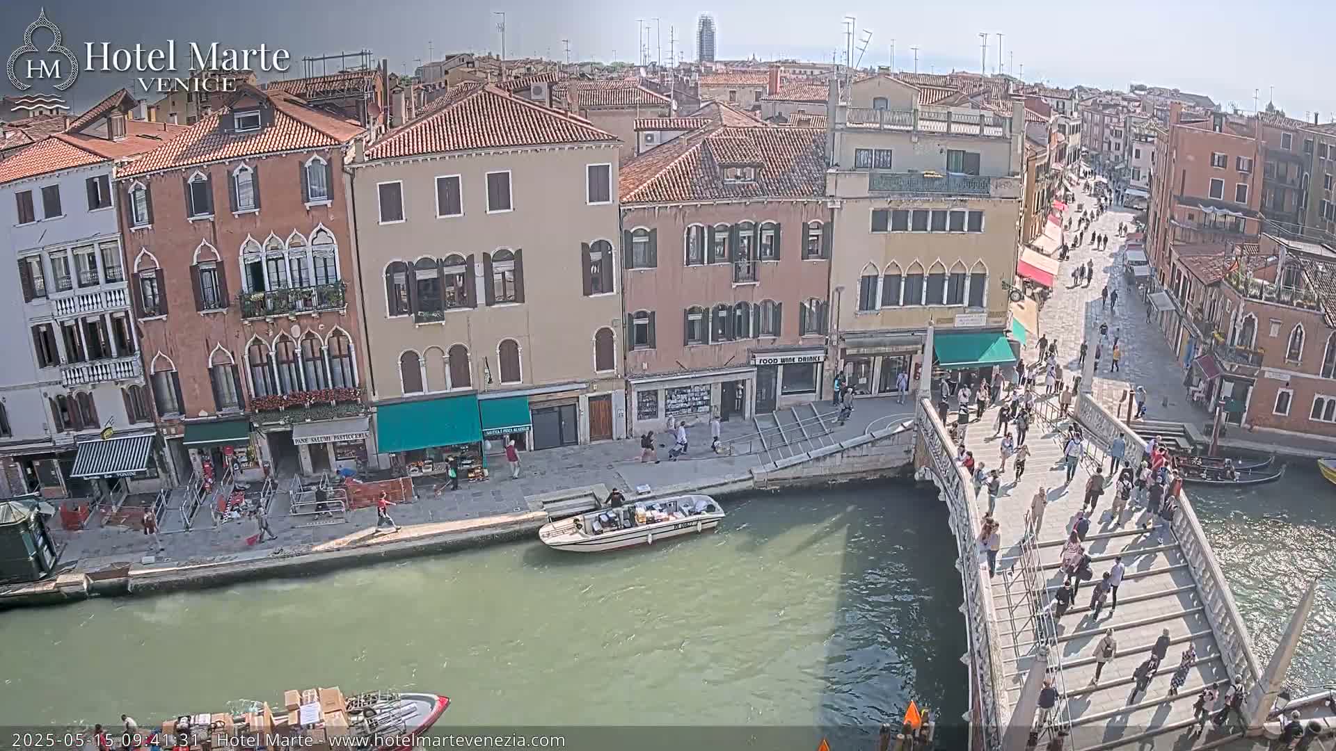 A sunny day in Venice shows a canal with a bridge crowded with pedestrians, boats on the water, and colorful buildings lining the waterway.