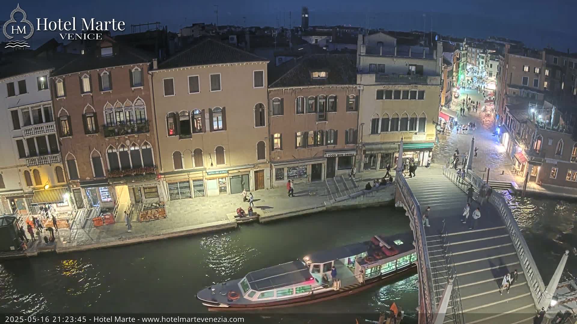 A nighttime view of a Venetian canal, showing a bridge with pedestrians, watercraft, and buildings lit along the waterway.