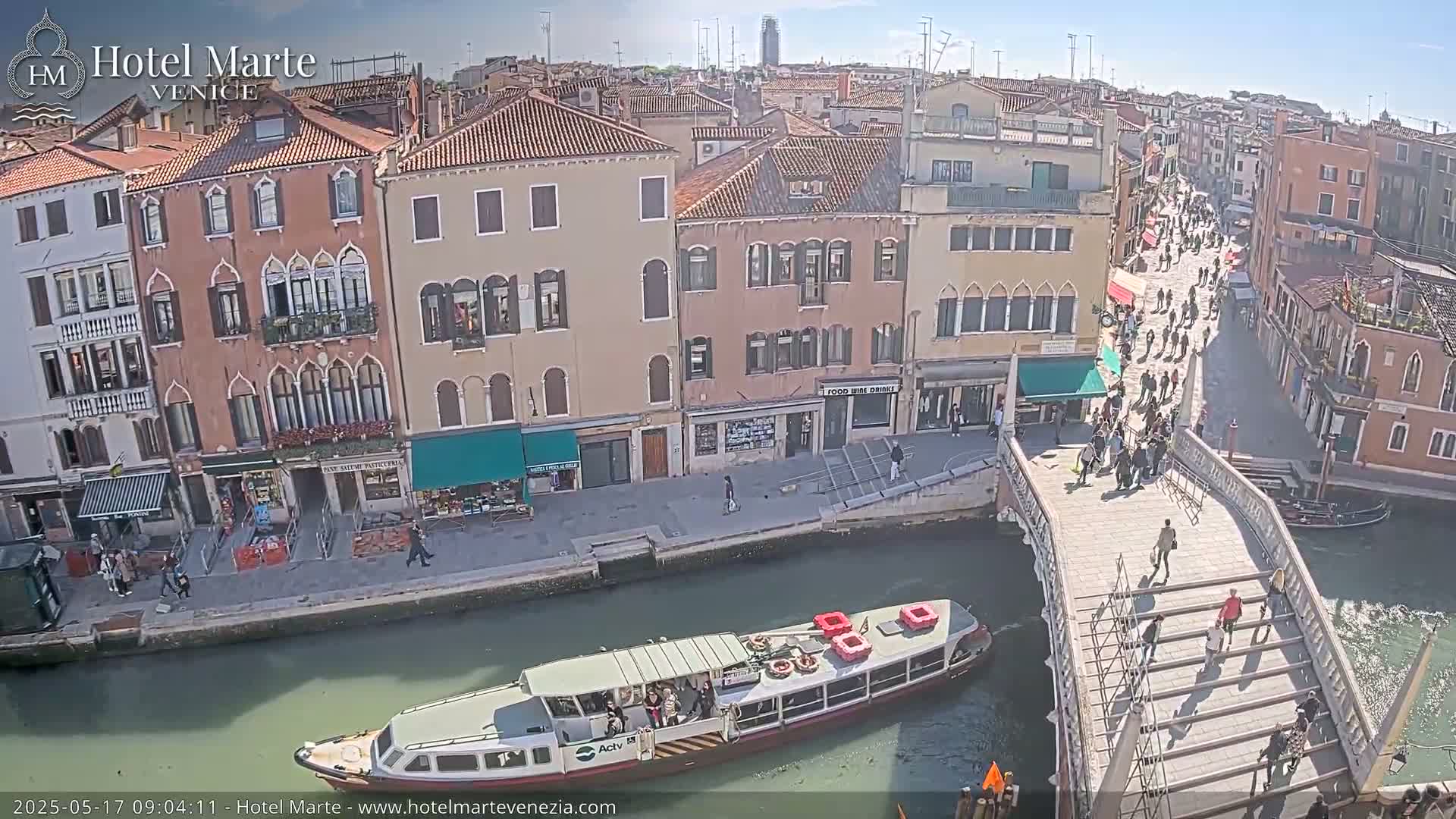 A sunny day in Venice shows a waterway with a boat, a bridge with pedestrians, and numerous buildings lining the canals.