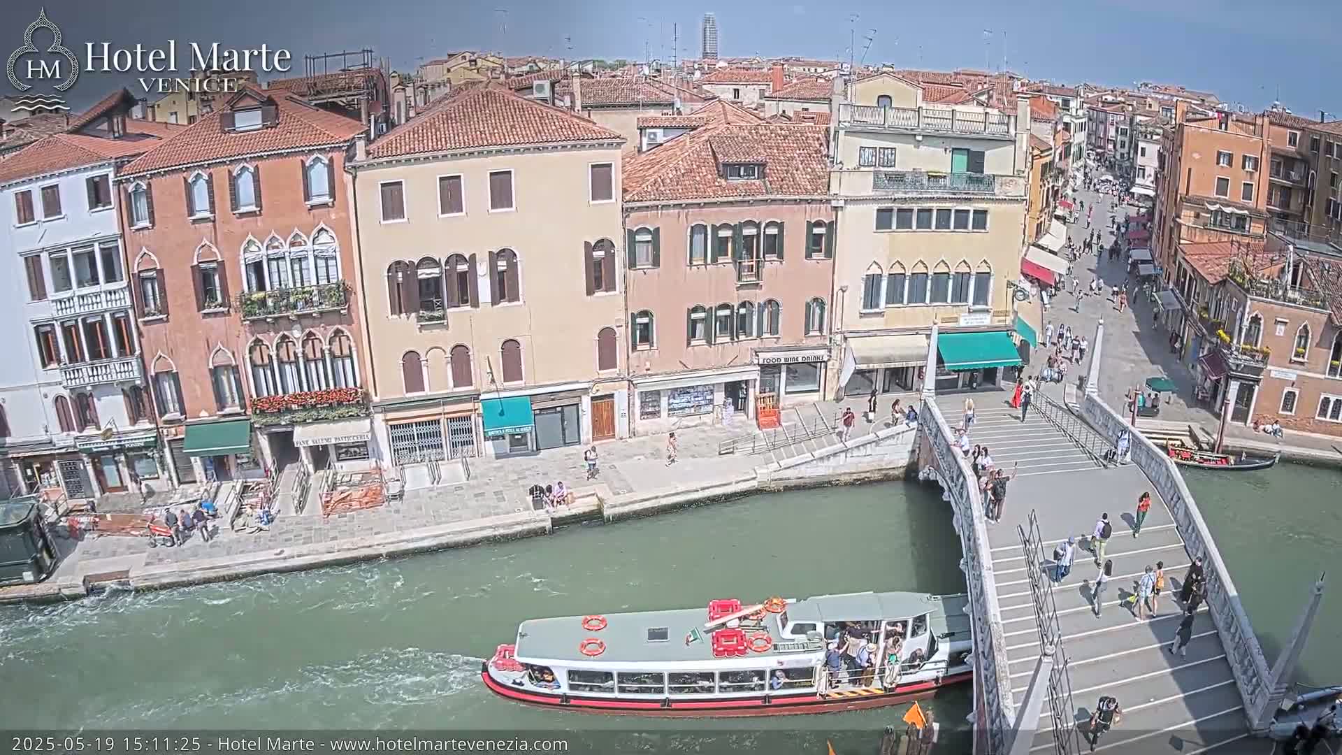 A sunny day in Venice shows a canal with a passenger boat and a bridge with people walking across it, alongside buildings lining the waterway.