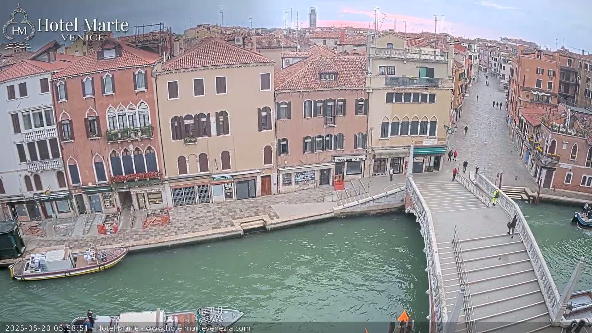A canal in Venice, with several buildings lining its sides, a bridge spanning the waterway, and a few pedestrians visible on the bridge and street, under an overcast sky.