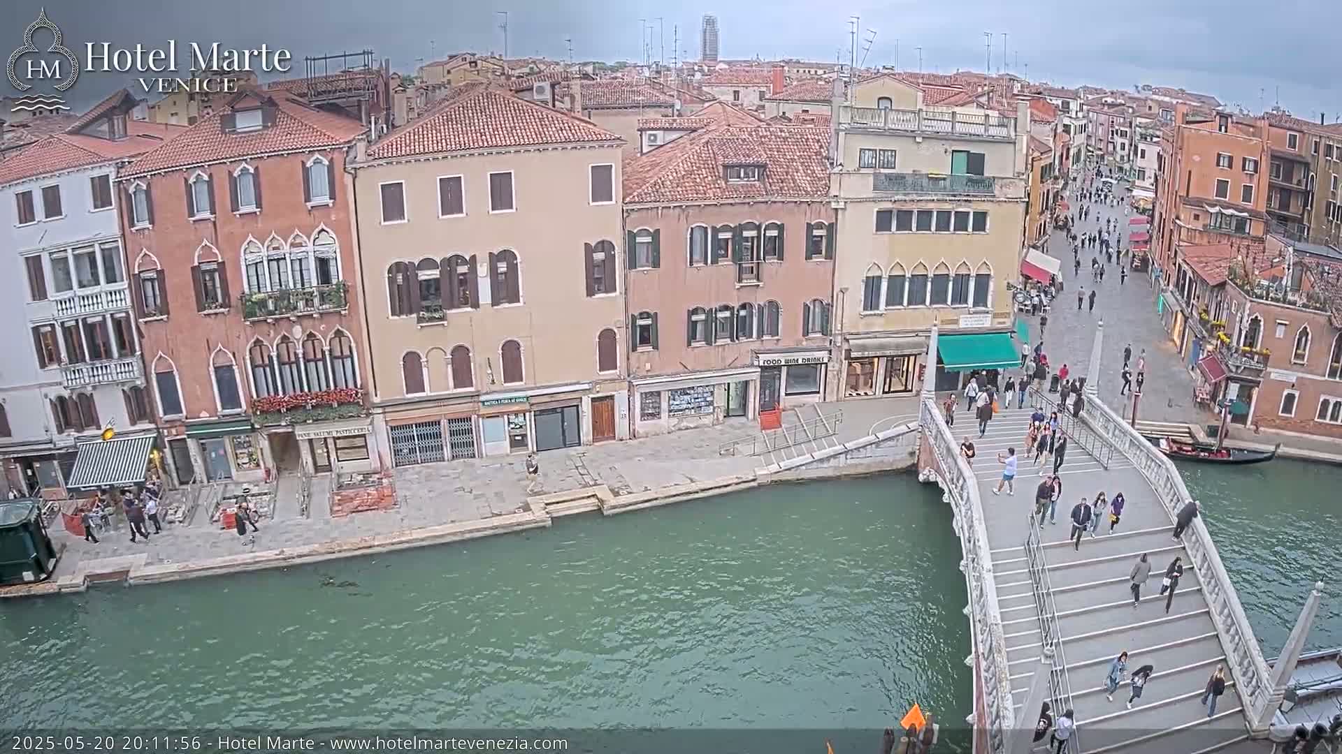 A canal in Venice, under an overcast sky, shows several buildings lining the waterway, with people walking across a bridge spanning the canal.