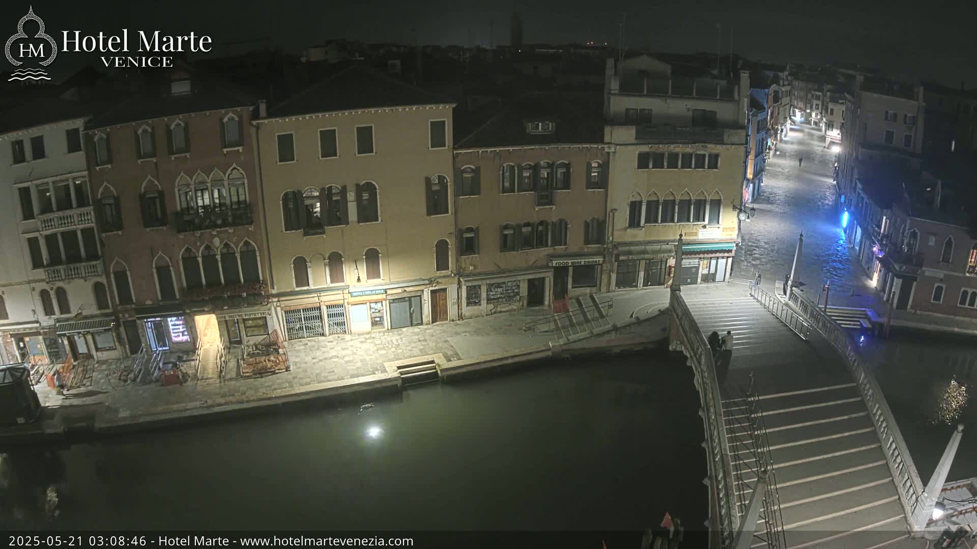A nighttime view of a canal in Venice, showing several buildings along the waterway and a bridge with people on it, under dark conditions.