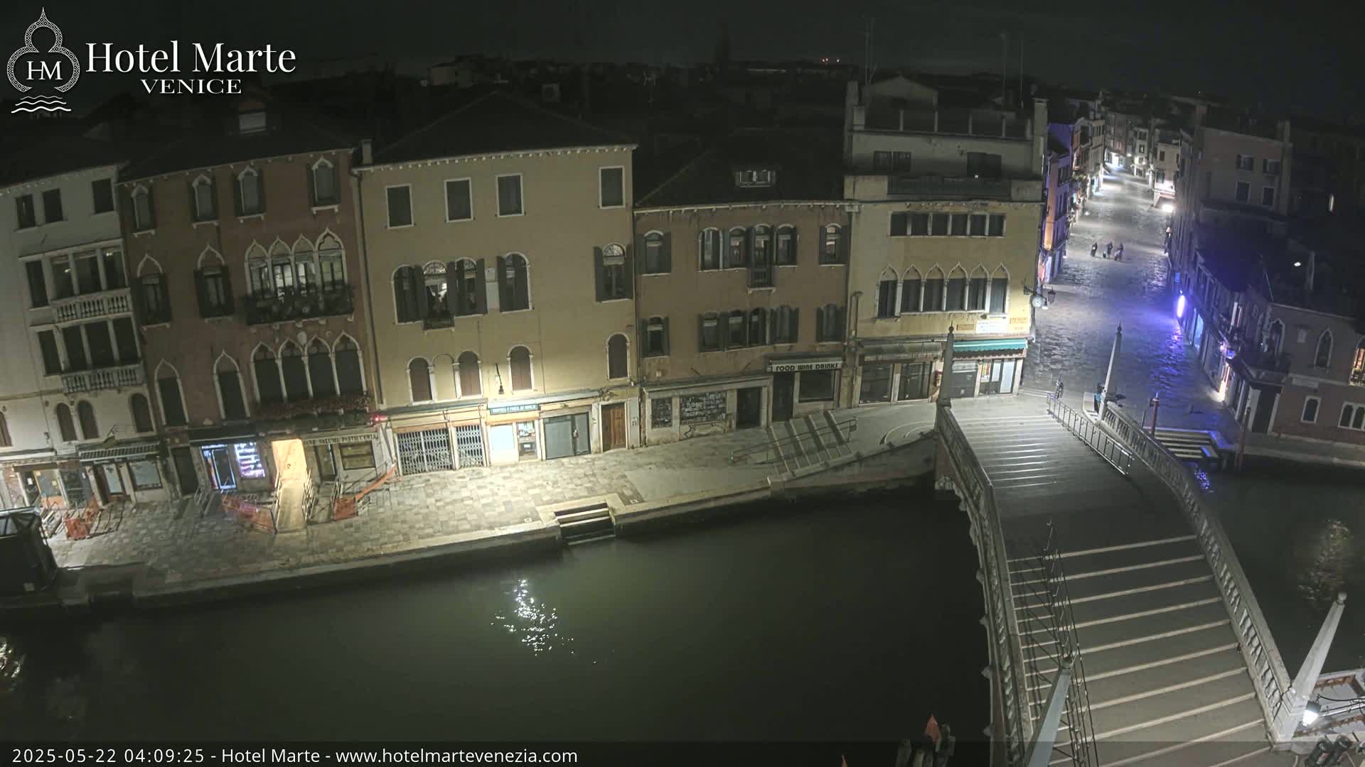 A nighttime view of a canal in Venice, showing several buildings lining the waterway and a stone bridge crossing the canal, with the water appearing calm.