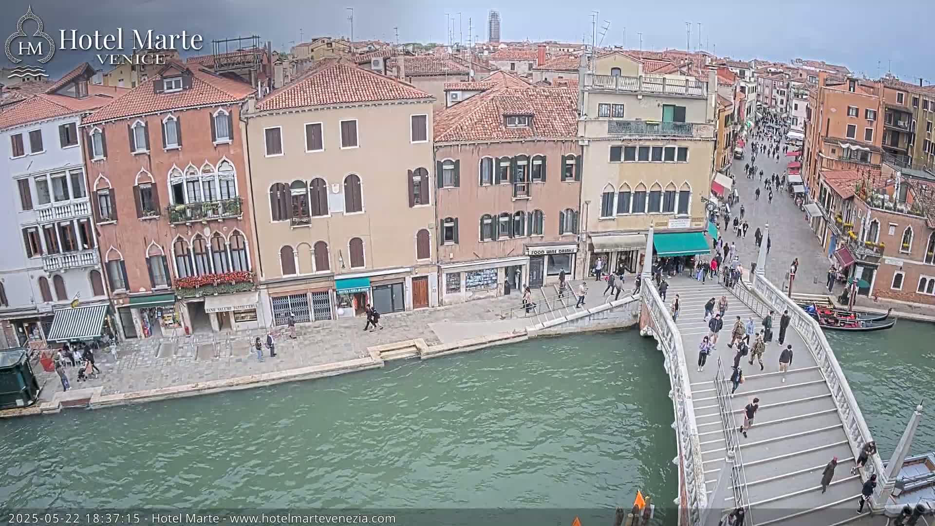 A canal in Venice, Italy, under an overcast sky, shows a bridge with pedestrians crossing it, and numerous buildings lining the waterway.