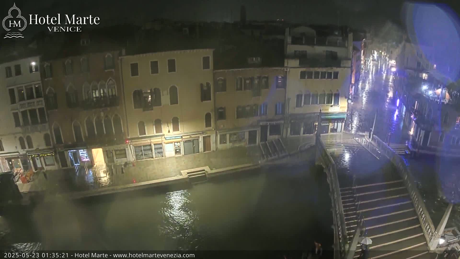 A nighttime view of a flooded canal in Venice, with rain falling and water covering a bridge and the street.