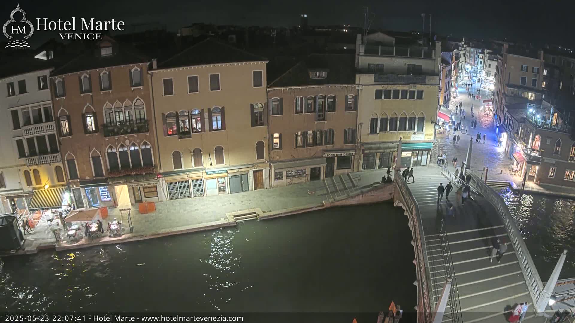 A nighttime view of a Venetian canal, showing buildings lining the waterway, pedestrians crossing a bridge, and several people sitting outside a restaurant.