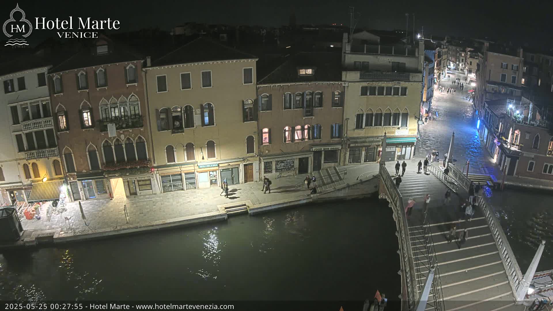 A nighttime view of a canal in Venice, Italy, shows several buildings along the water's edge and pedestrians crossing a bridge.