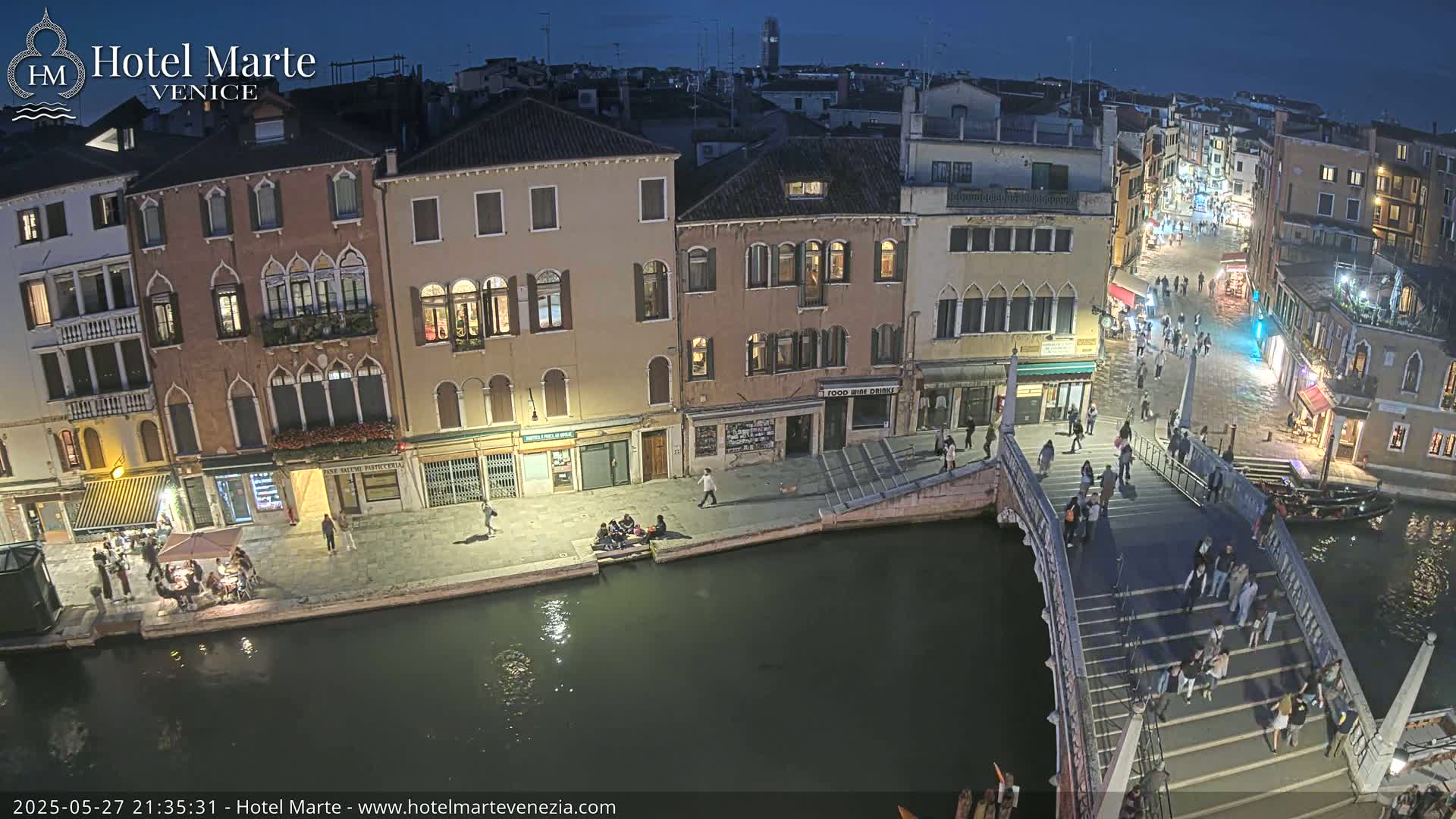 A nighttime view of a Venetian canal, showing pedestrians crossing a bridge and strolling along the water's edge, with buildings lining the canal.