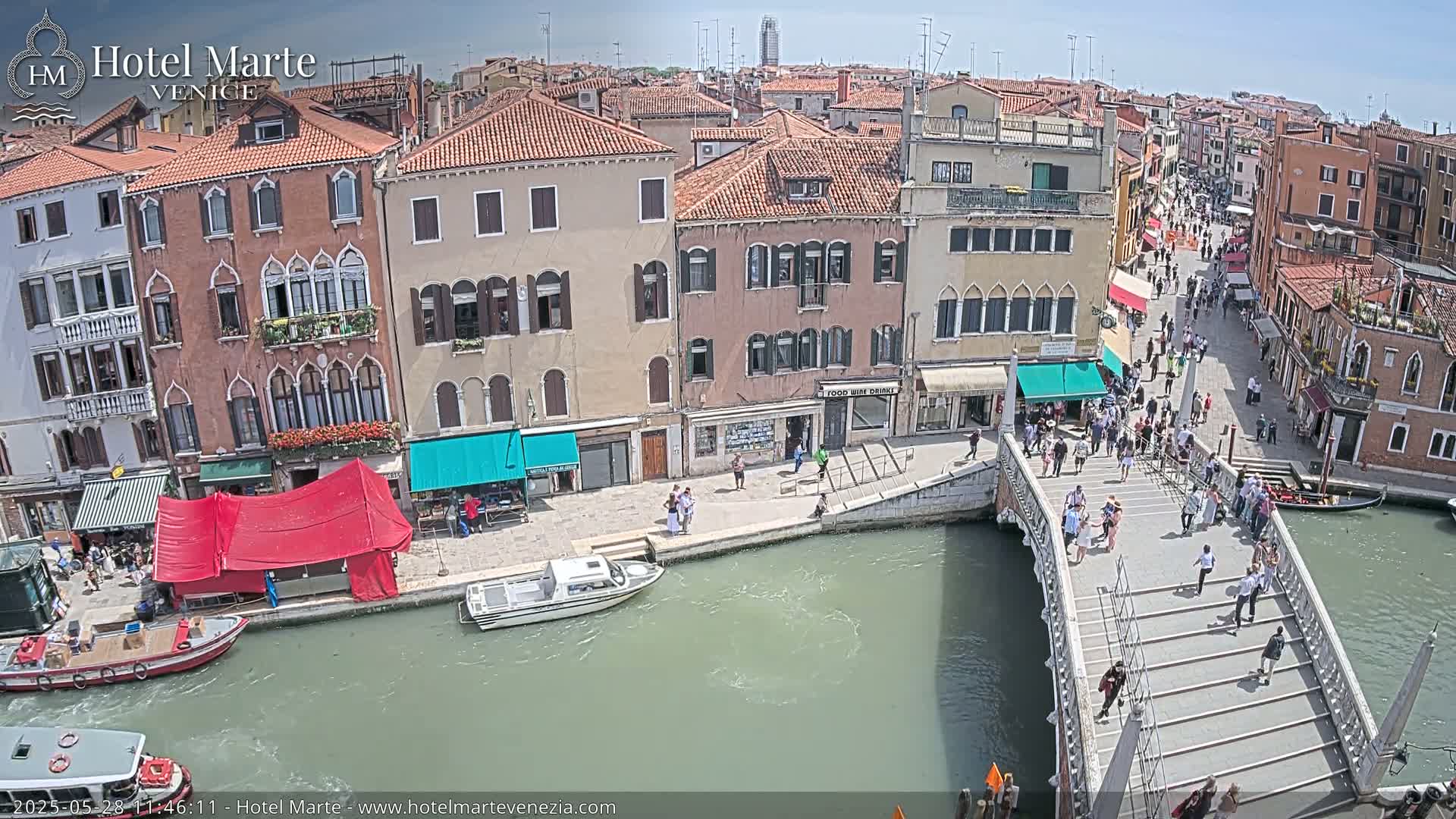 A sunny day in Venice shows a canal with boats, pedestrians crossing a bridge, and colorful buildings lining the waterway.