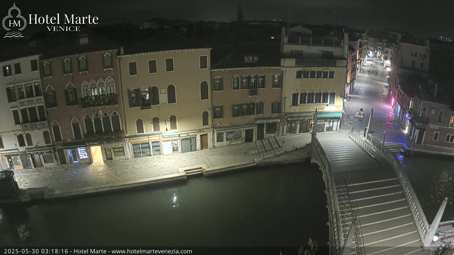A nighttime view of a canal in Venice, showing several buildings along the waterway and a bridge with steps leading down to the water's edge.