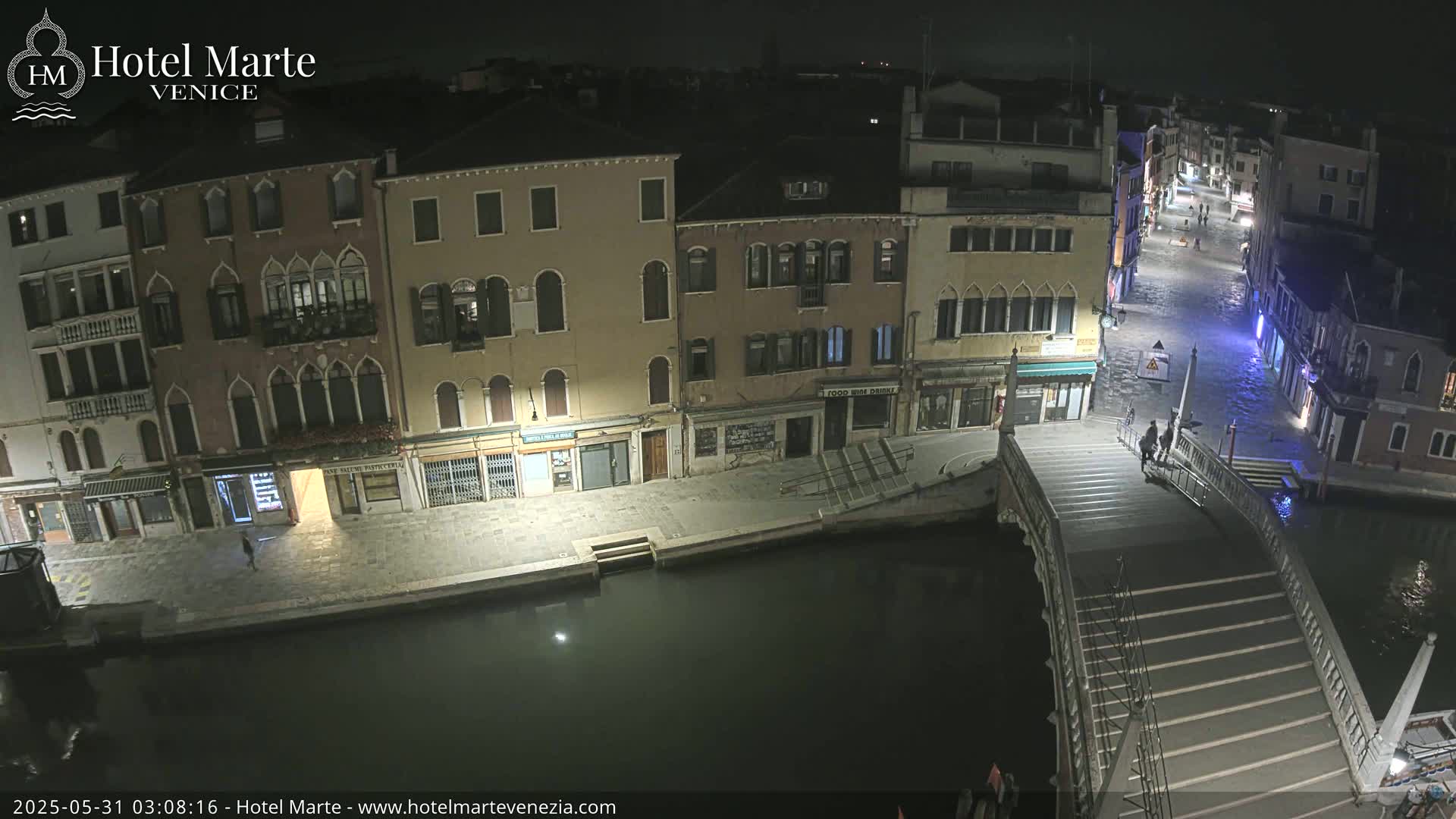 A nighttime view of a canal in Venice, showing several buildings along the waterway and a bridge with a few pedestrians.