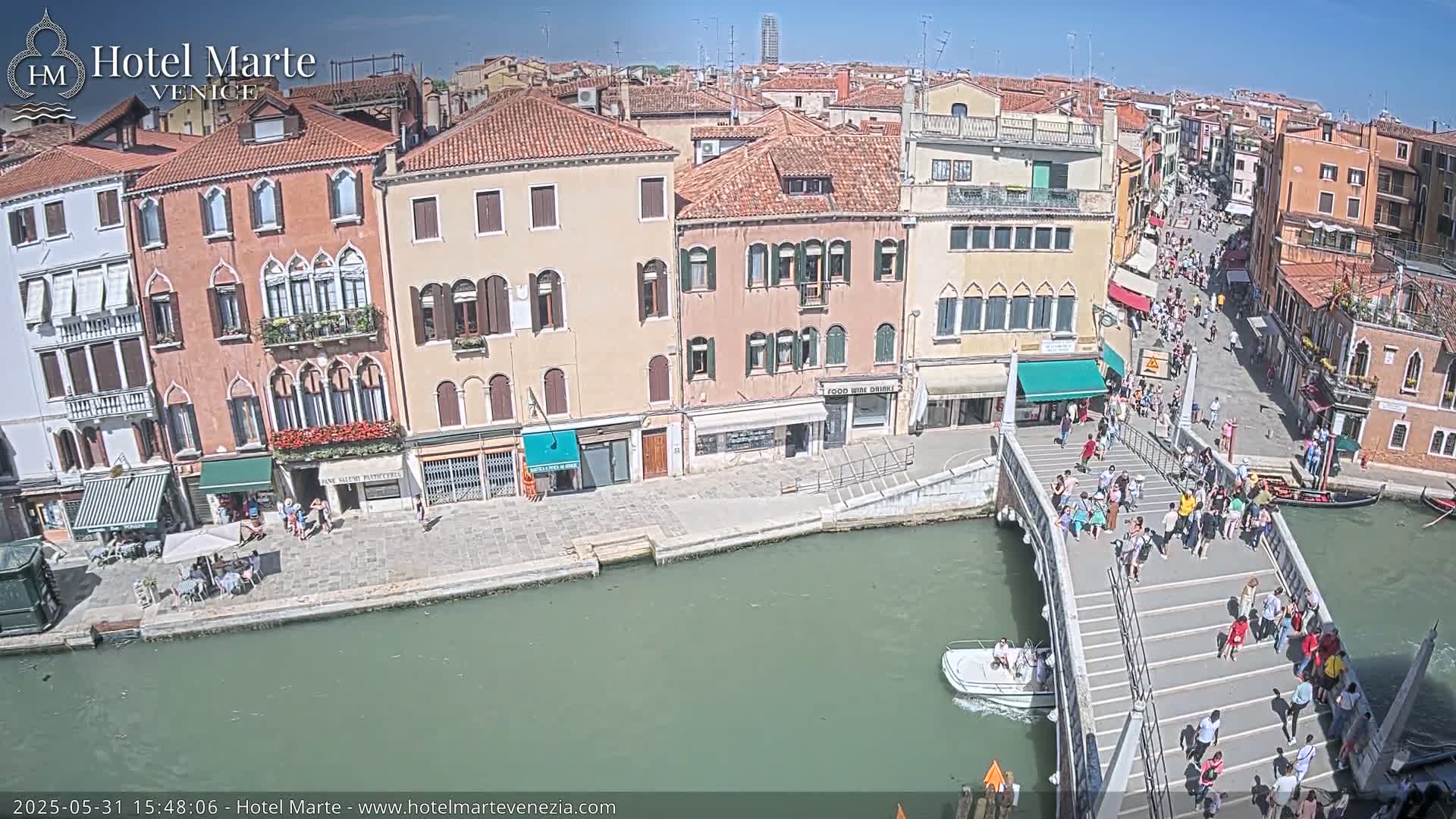 A sunny day in Venice shows a canal with a footbridge crowded with people, boats, and buildings lining the waterway.