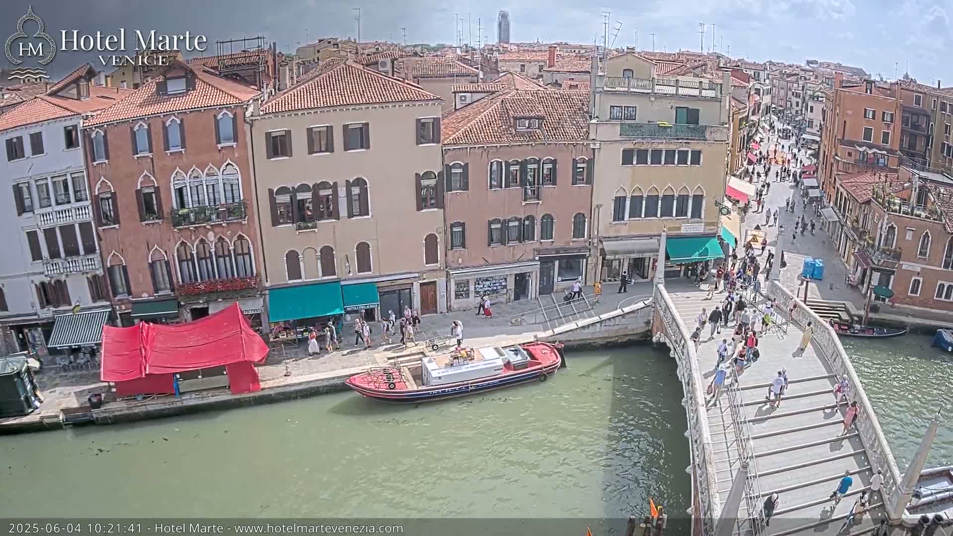 A sunny day in Venice shows a canal with a bridge crowded with people, boats, and colorful buildings lining the waterway.