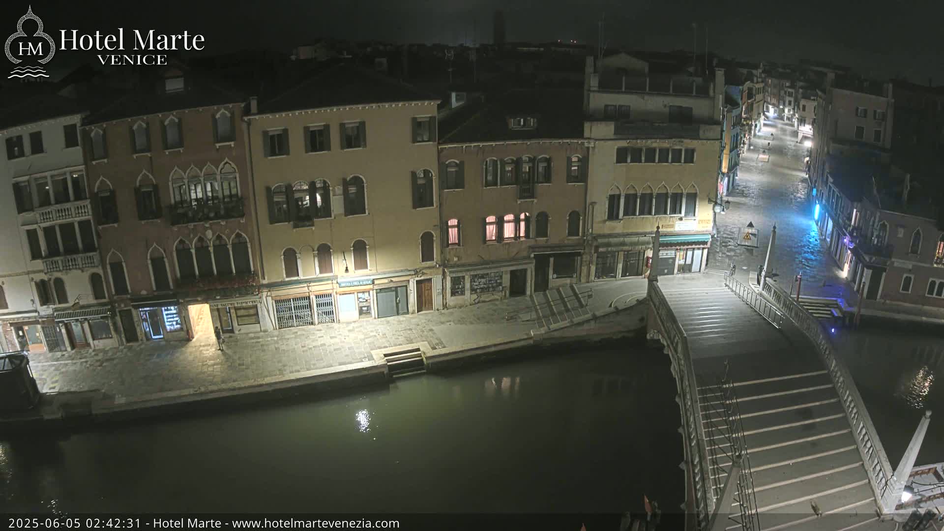 A nighttime view of a canal in Venice, showing several buildings along the waterway, a bridge, and water levels appearing slightly elevated.