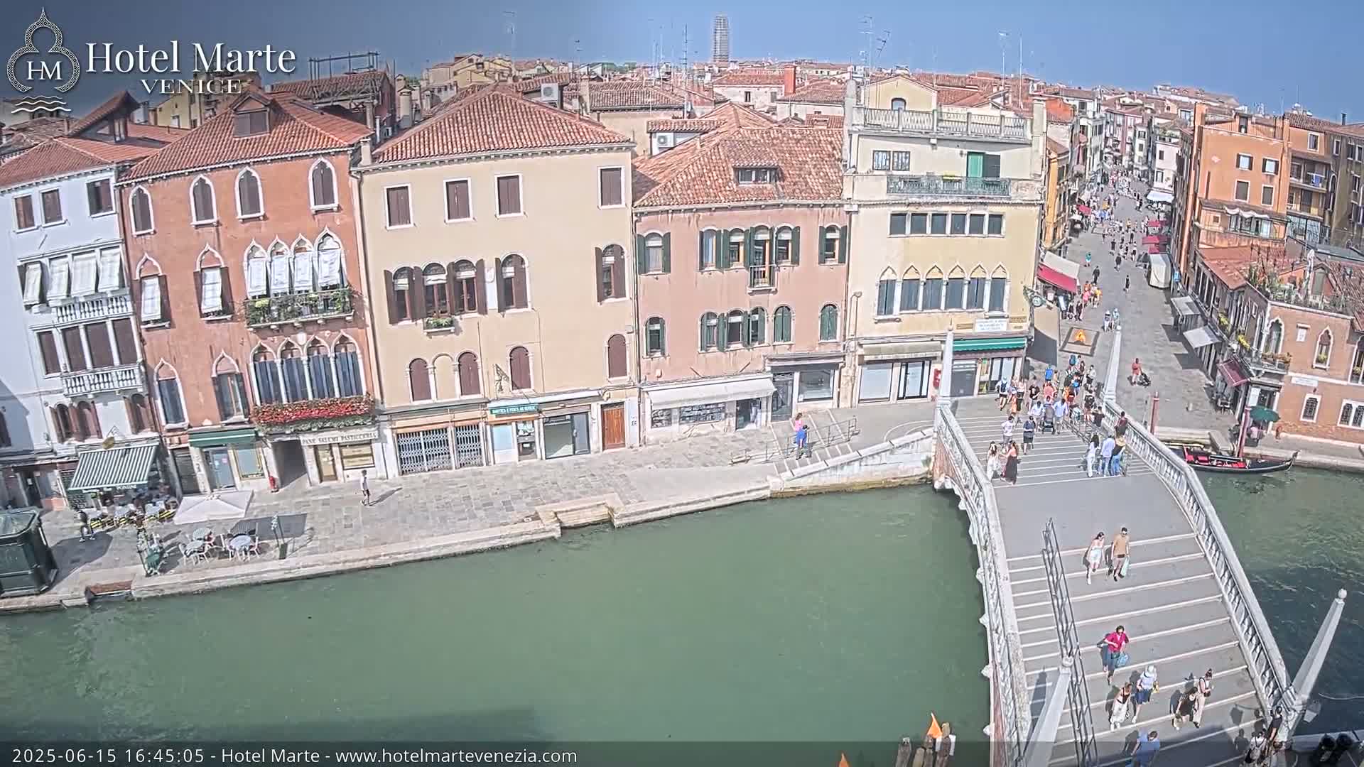 A sunny day in Venice shows a canal with a footbridge crowded with people,  lined with buildings along its sides.