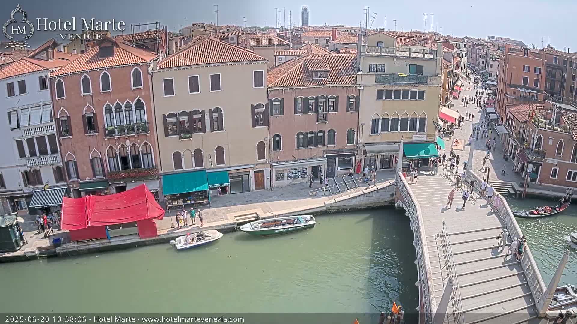 A sunny day in Venice shows a canal with gondolas, a bridge with pedestrians, and colorful buildings lining the waterway;  a red market stall is visible near the water's edge.