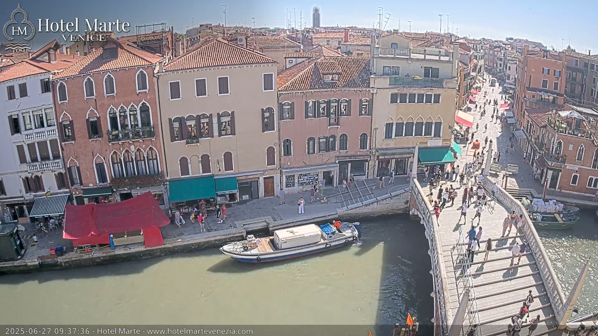 A sunny day in Venice shows a canal with a bridge crowded with pedestrians, boats on the water, and colorful buildings lining the waterway.