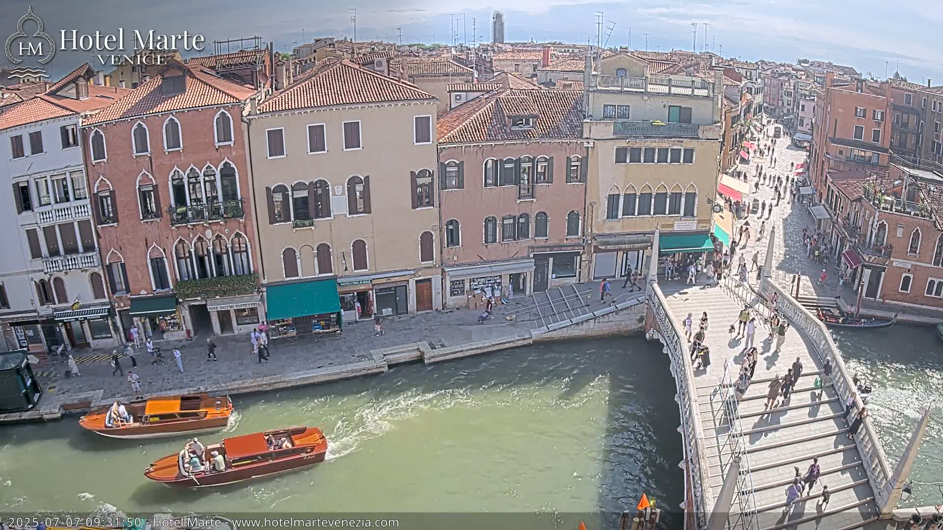 A sunny day in Venice shows a canal with boats, pedestrians on a bridge and buildings lining the waterway.