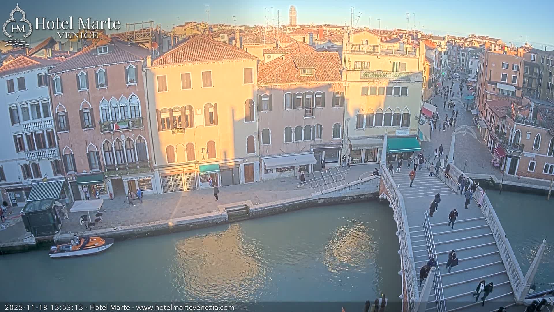 On an overcast day, a bustling canal city scene features a crowded stone bridge over a teal canal, with numerous pedestrians walking along adjacent streets lined with multi-story historic buildings, and a small boat docked along the waterway.