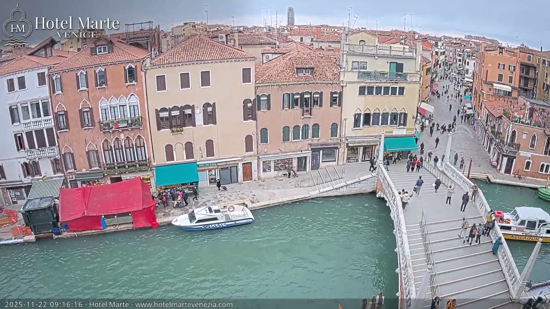 A bustling view of Venice on an overcast day shows people crossing a large pedestrian bridge over a teal-colored canal, flanked by historic multi-story buildings and a police boat navigating the waterway.