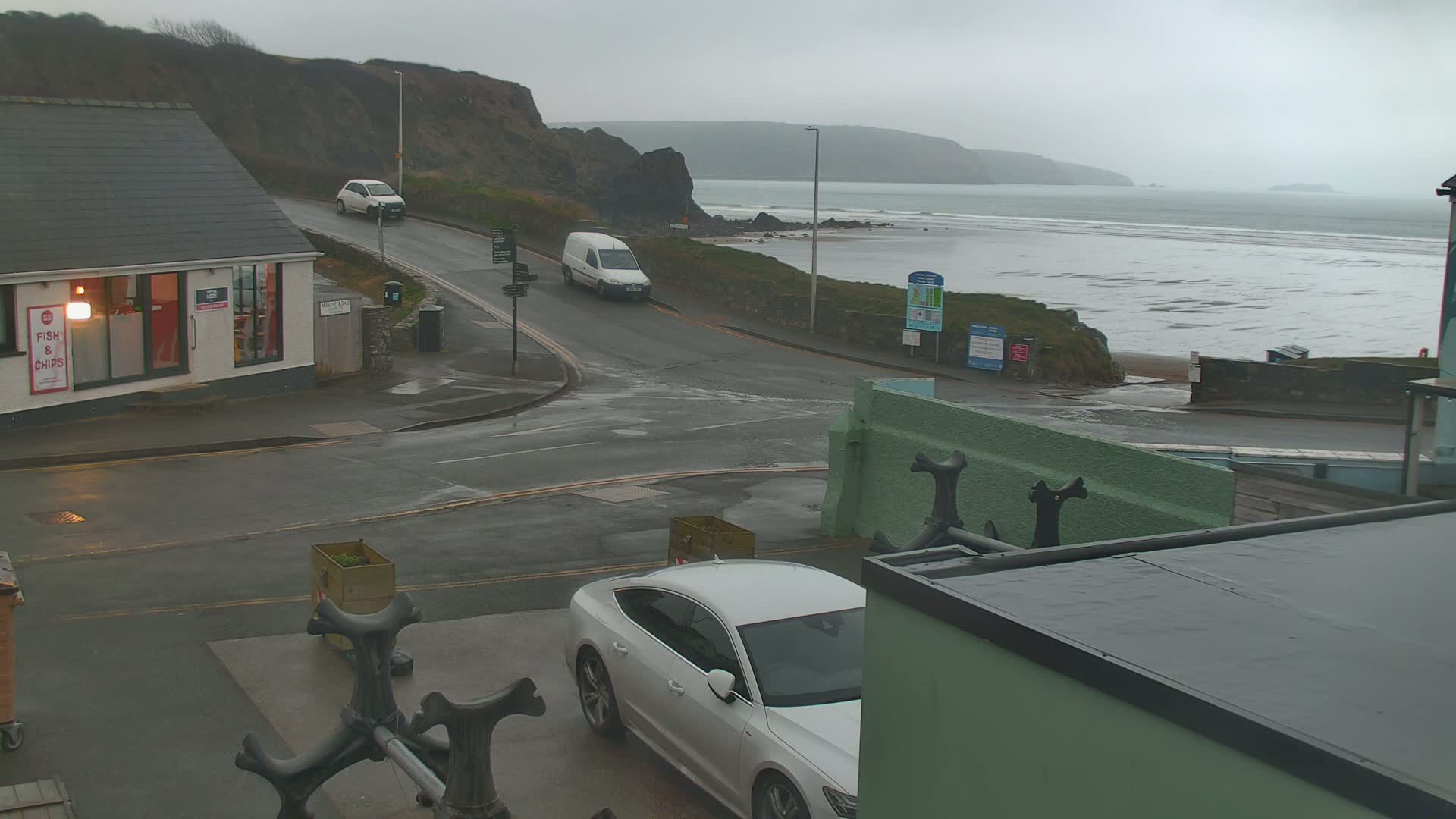 A wet, overcast day at a seaside town shows a road intersection near a fish and chips shop, with cars parked and driving, and a view of a beach and ocean in the background.