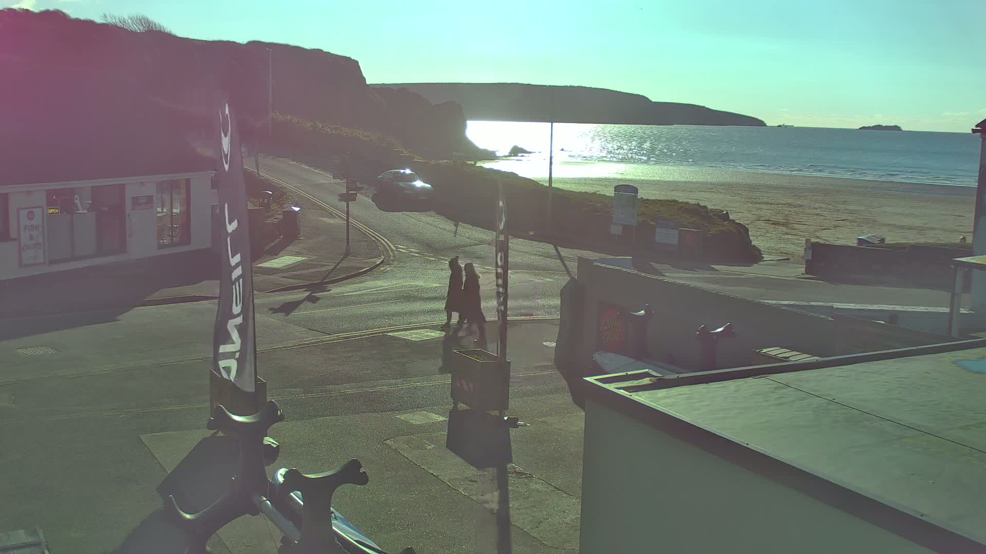 A sunny day at a coastal town shows a sandy beach, calm sea reflecting sunlight, a road intersection with pedestrians and a car, and buildings near the shore.