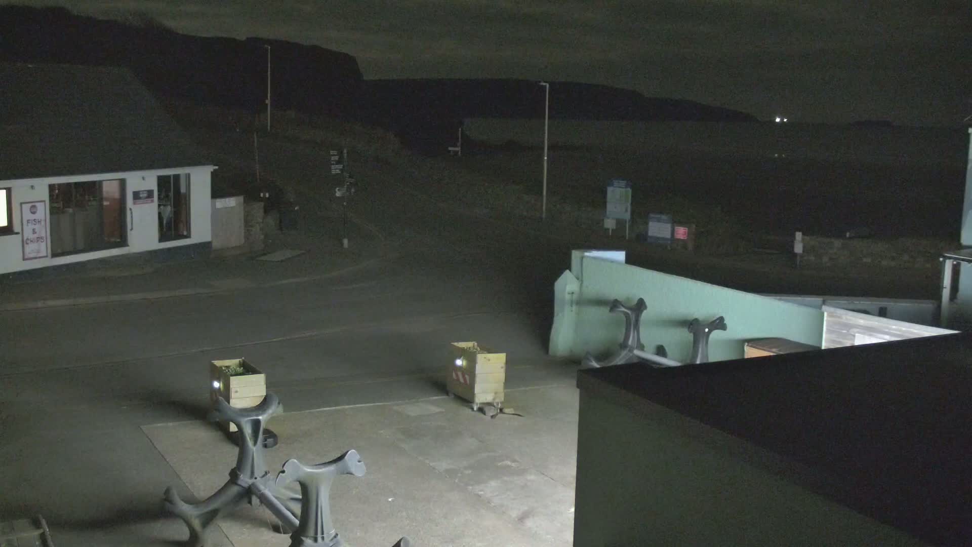 At night, a mostly empty paved area near a building and the coast is dimly lit, with two boxes and several dark, metal objects present.
