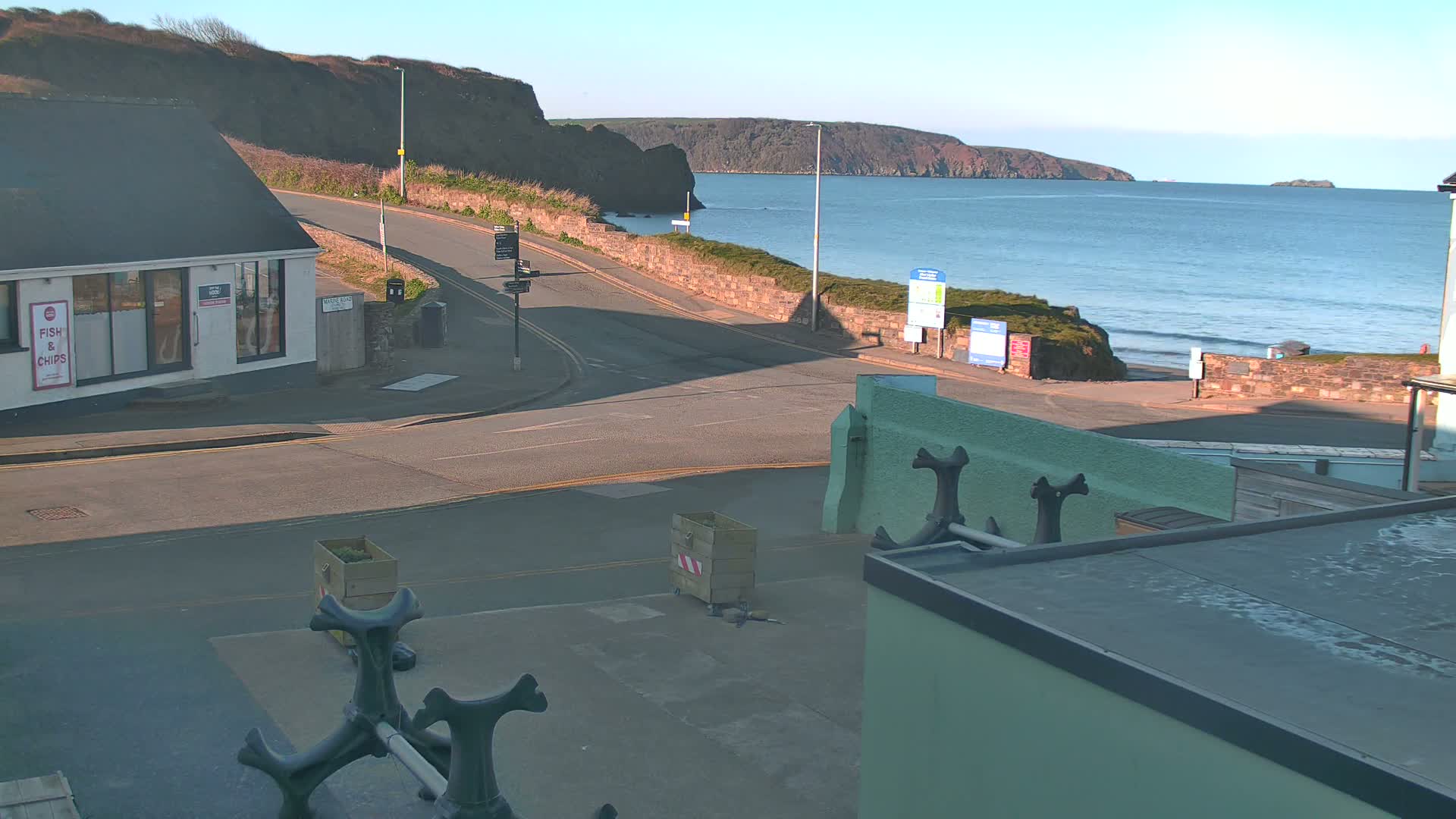 A sunny coastal scene shows a road curving toward a beach with calm ocean waves, beyond which lies a rocky coastline.