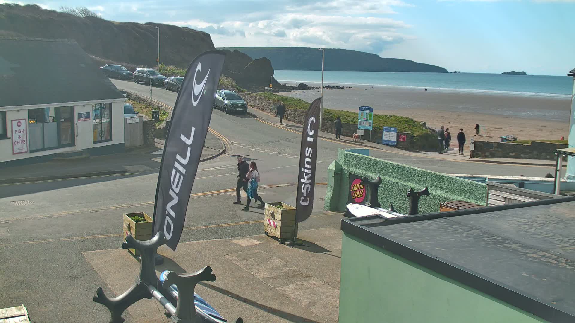 A sunny day at a beach town shows a sandy beach, ocean, and people walking along a road near buildings under a partly cloudy sky.