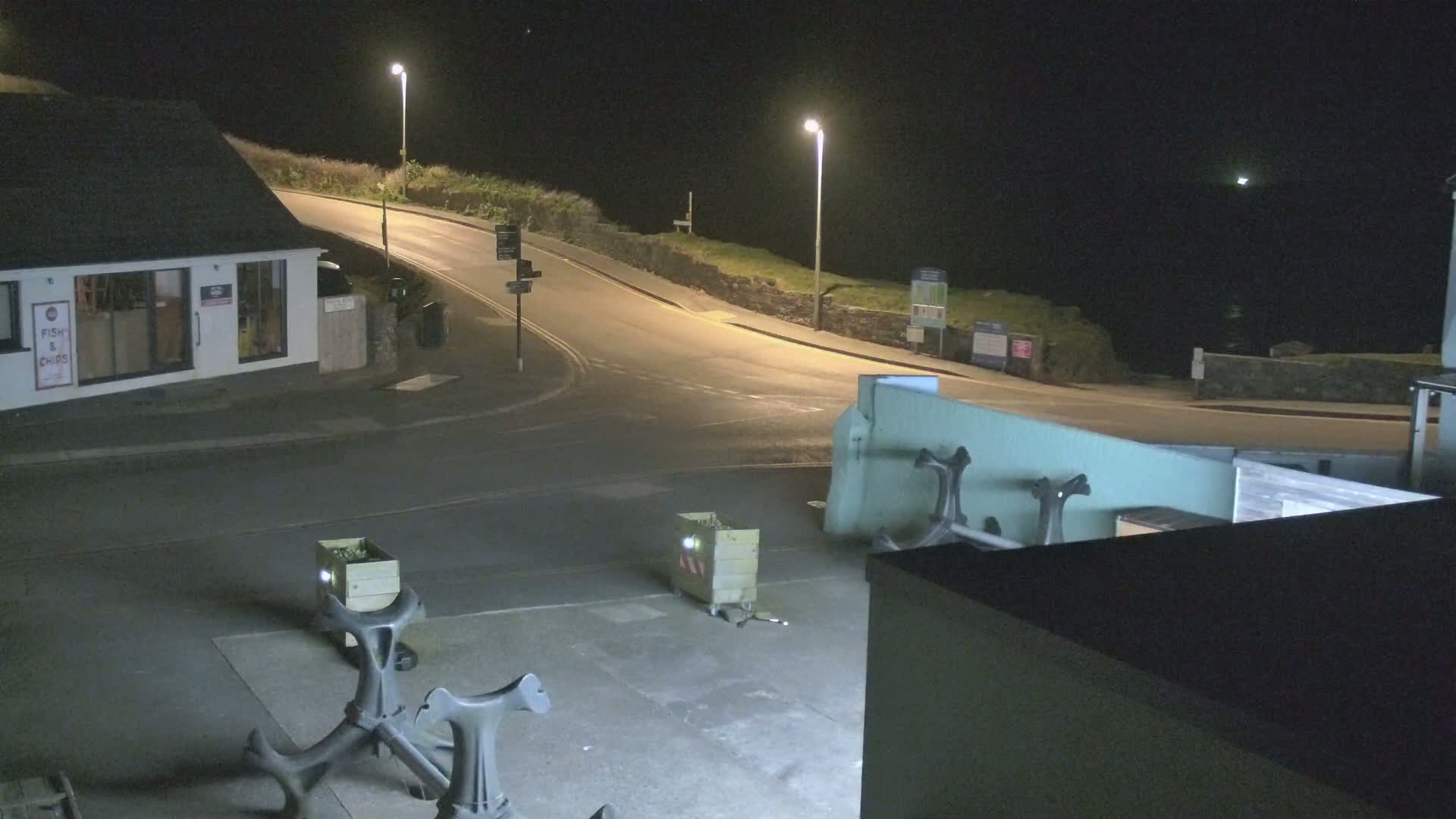 A nighttime view of a street corner near the ocean, illuminated by streetlights, showing a building, road, and several anchors on the ground.